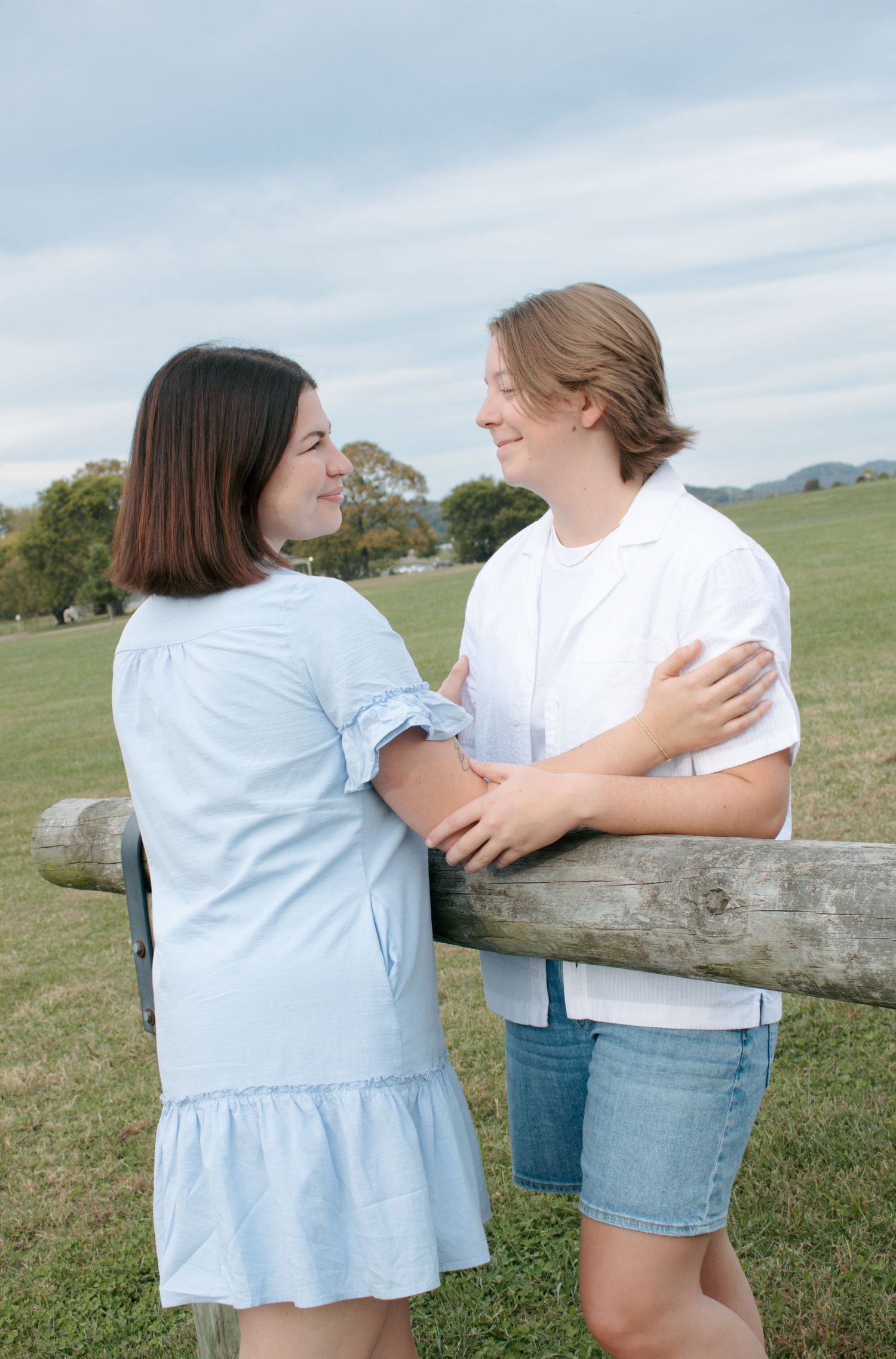 Two people in casual attire leaning on wooden fence in pastoral setting.
