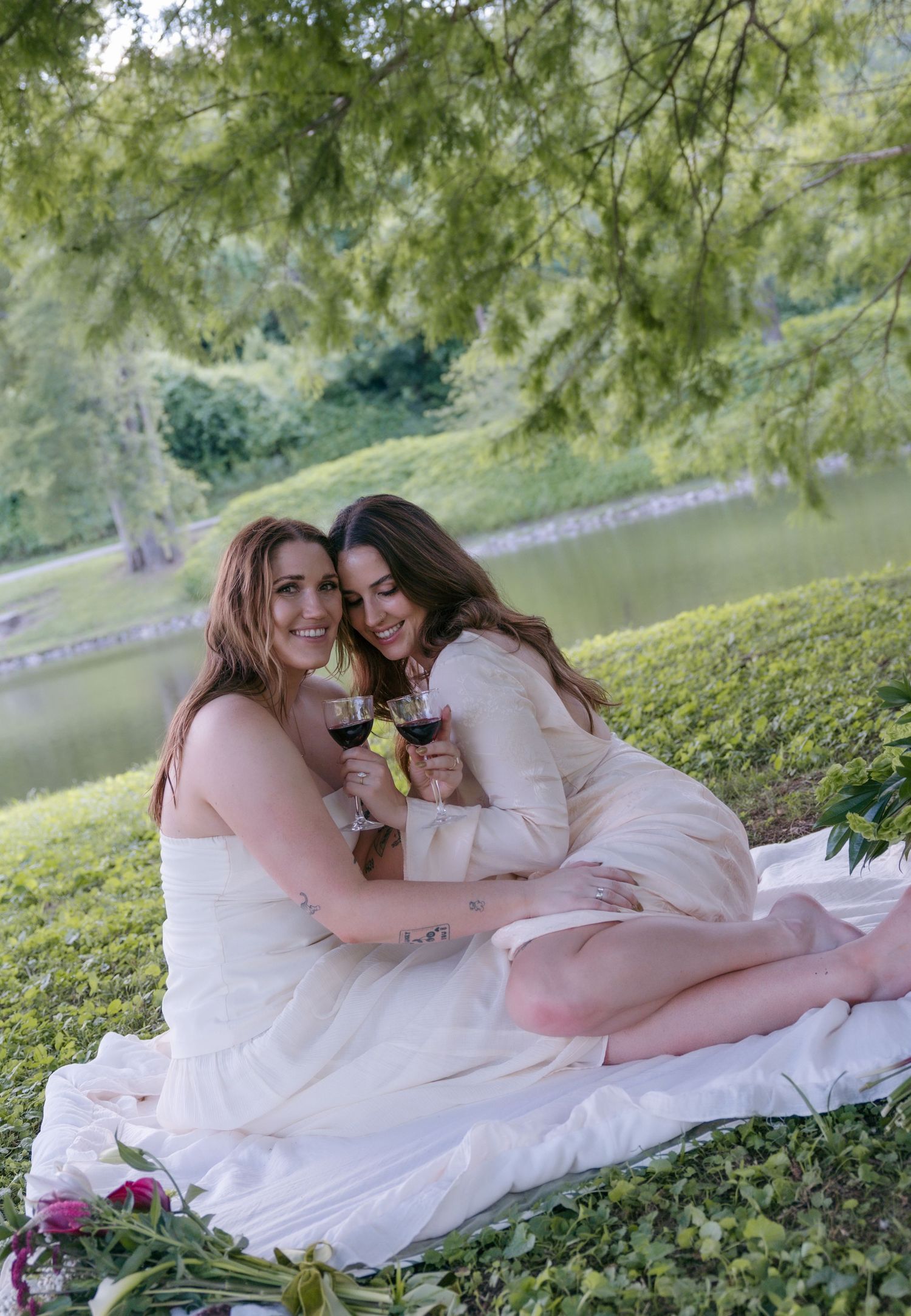 Two women in elegant dresses toasting wine glasses during a romantic outdoor picnic on a blanket by a serene lake.