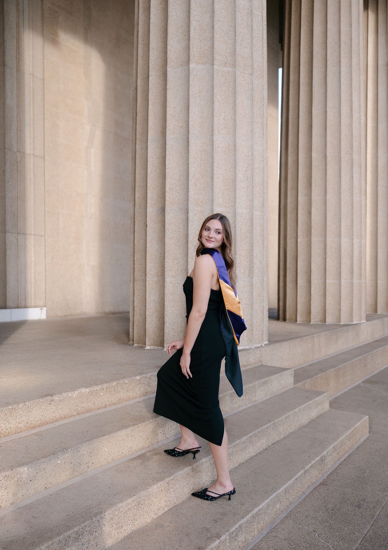 Young woman in black dress holding graduation regalia, posing on steps between large stone columns.