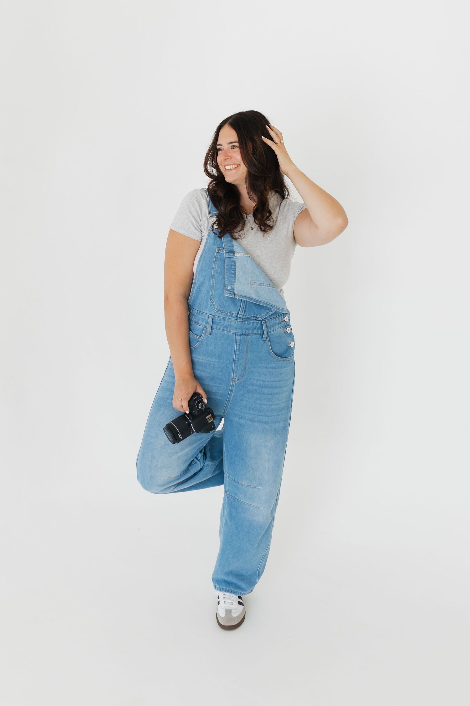Creative portrait of photographer in overalls posing with camera against white wall.