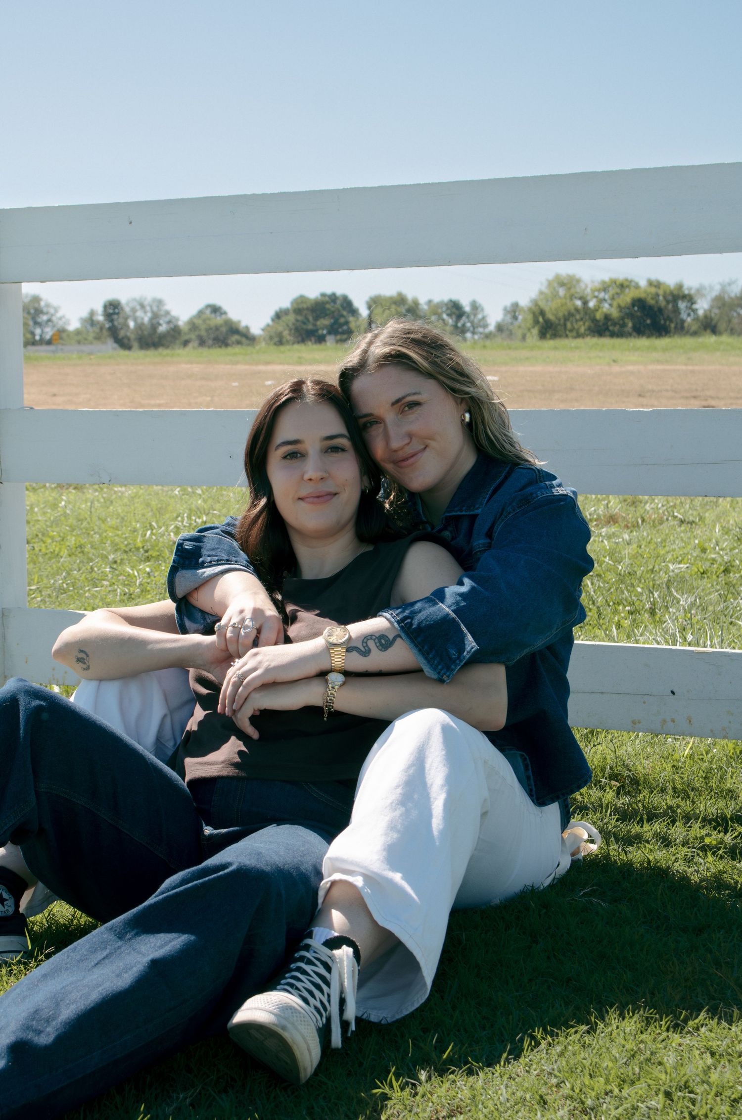 Two people sitting together on grass by a white fence on a sunny day.