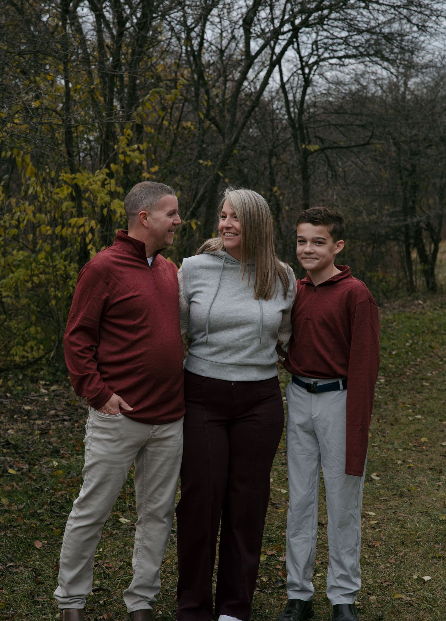 Family of three wearing burgundy and gray outfits poses together in a fall outdoor setting with bare trees in background.