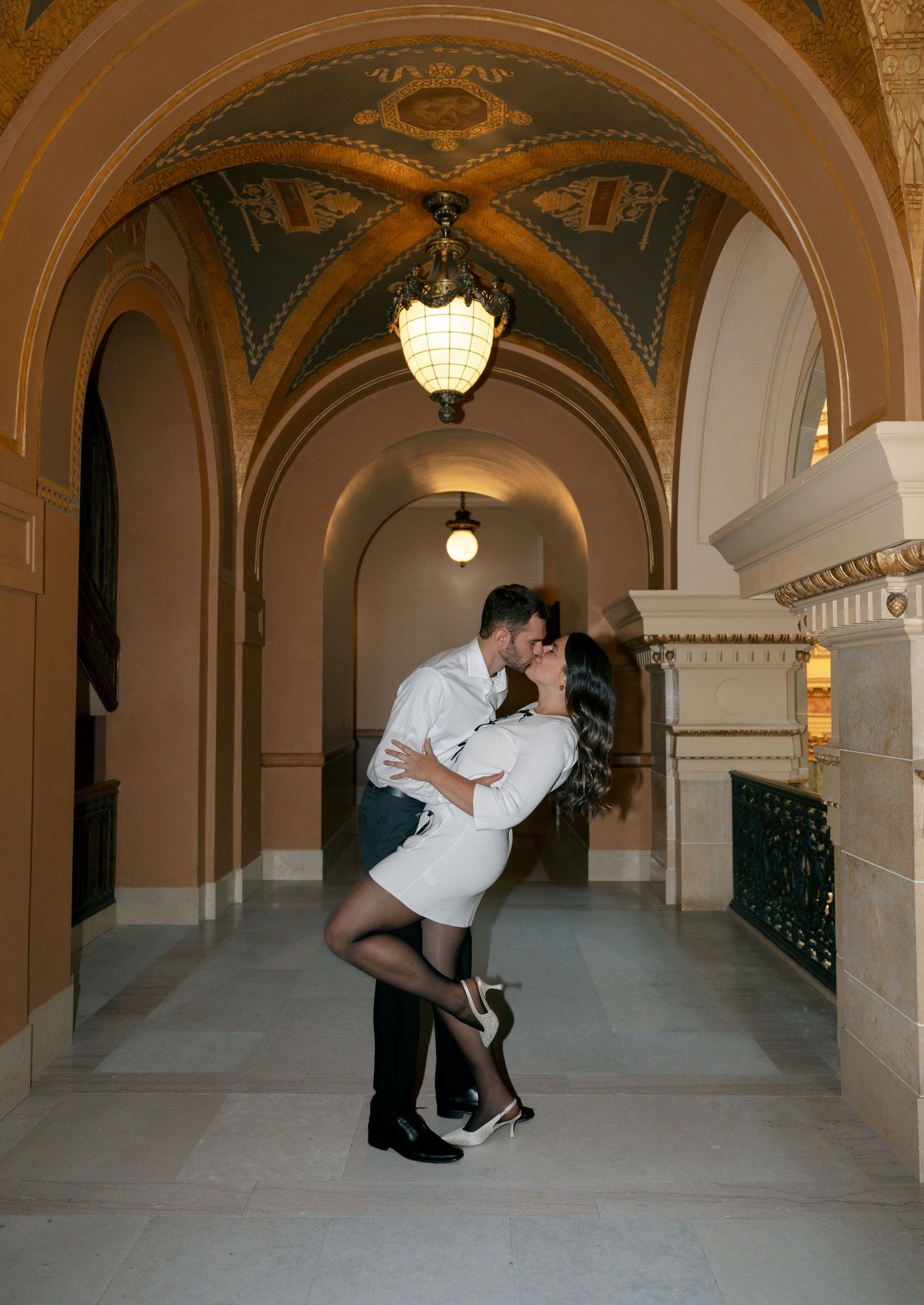 A romantic couple shares a passionate dip-style dance pose in an elegant arched hallway with ornate architectural details.