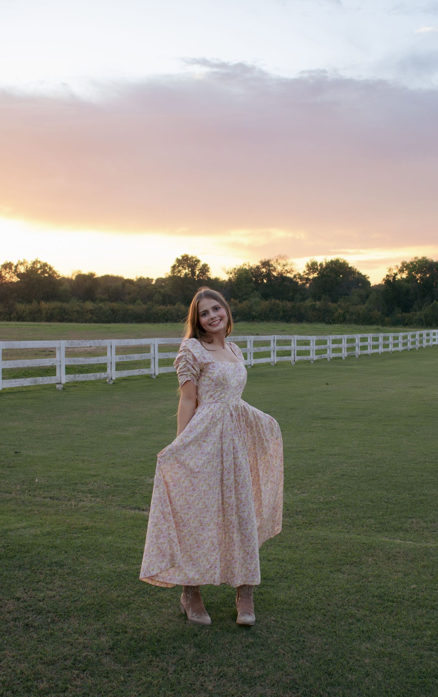A person in a floral dress twirls in a field with white fences at sunset.