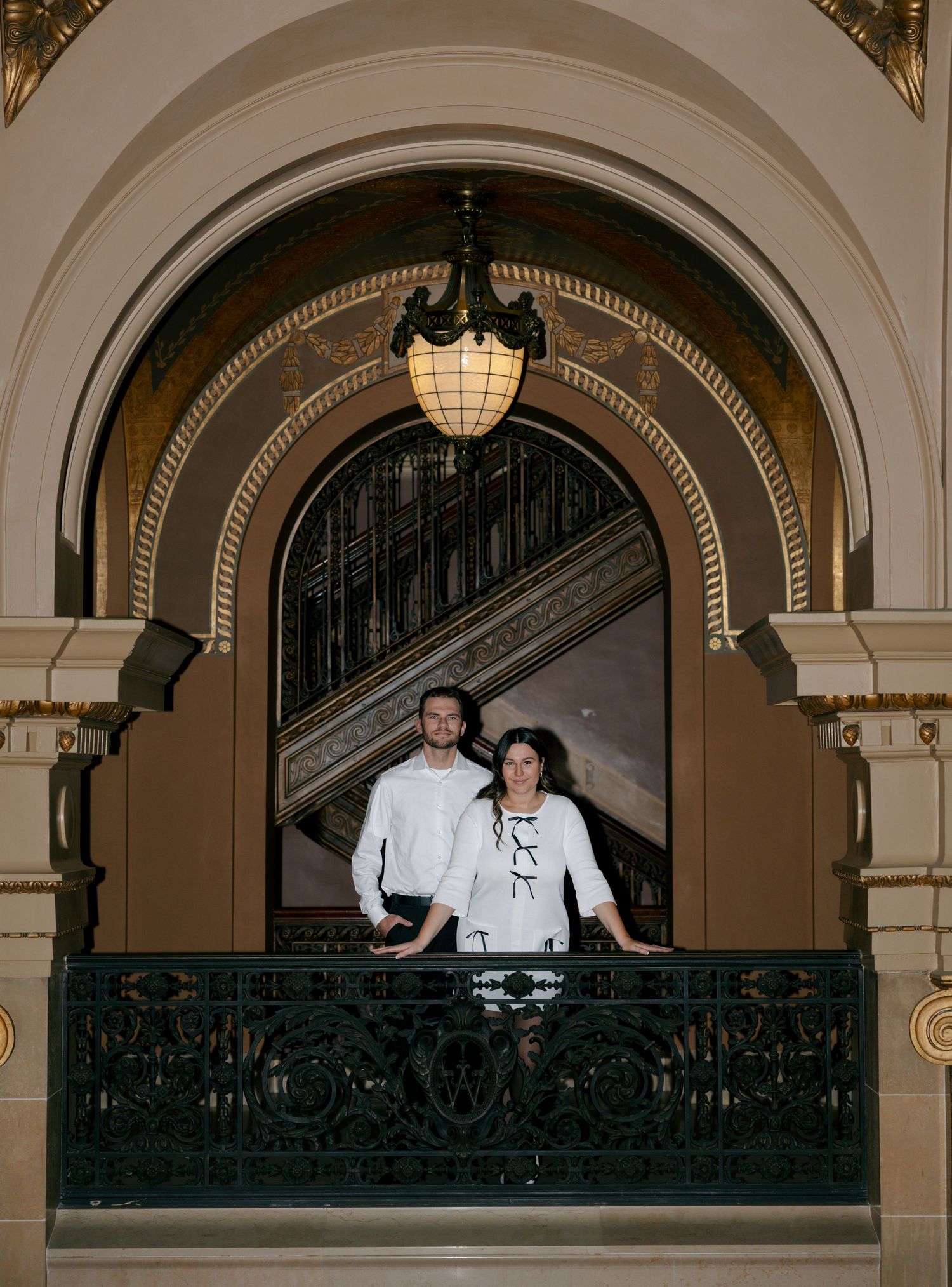 Two chefs in white uniforms converse on an elegant balcony with ornate architectural details and arched doorways.