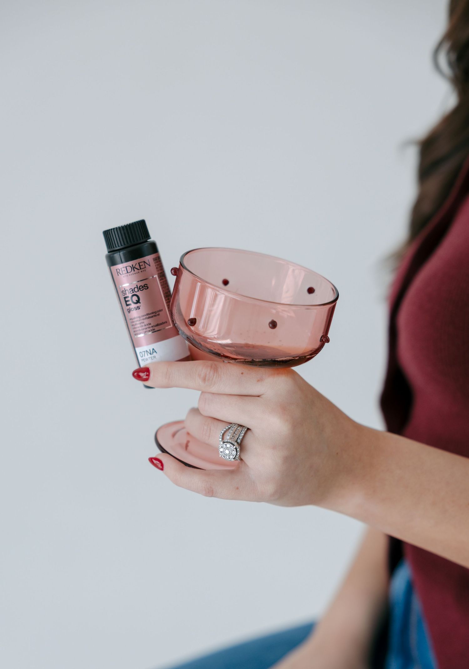 Woman holding a pink salon bowl and Redken hair color bottle, wearing a diamond ring with red nail polish.