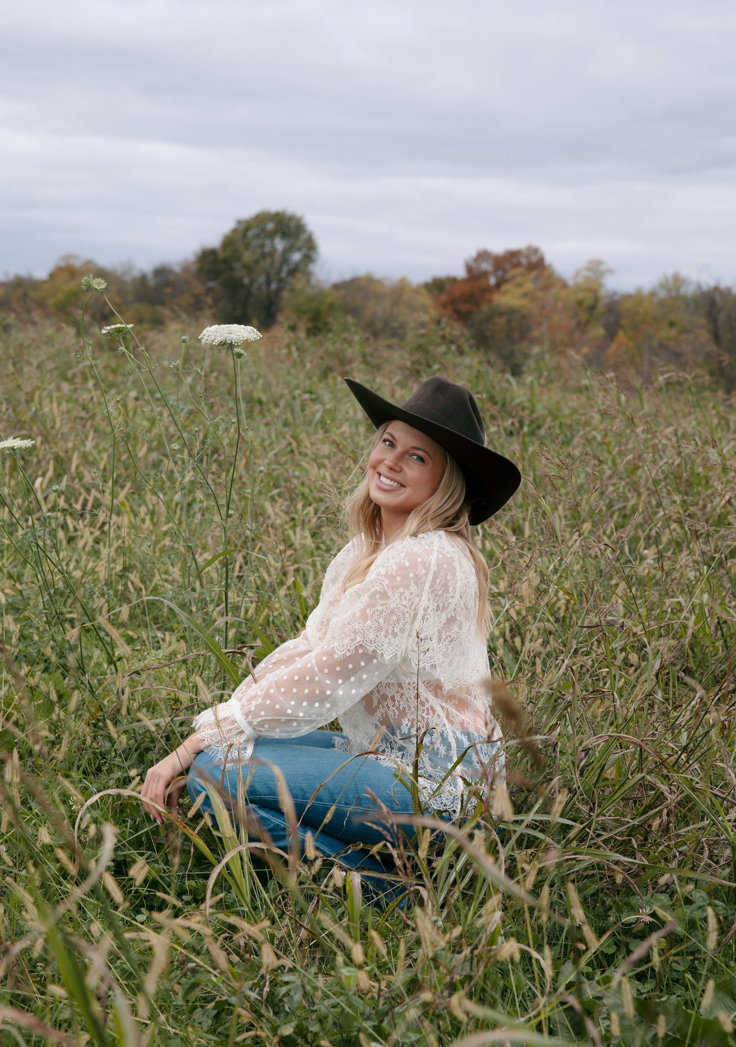 A person in a black hat and lace top reclines in a grassy autumn field during an outdoor photoshoot.