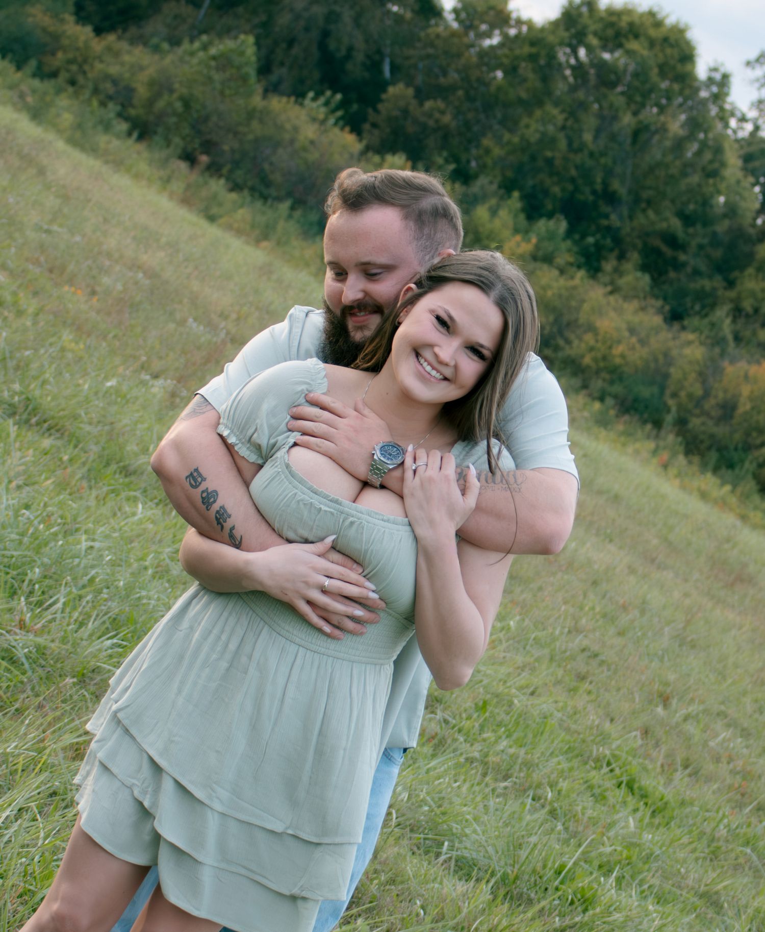 Couple sharing playful moment in sage green outfits on grassy hillside.
