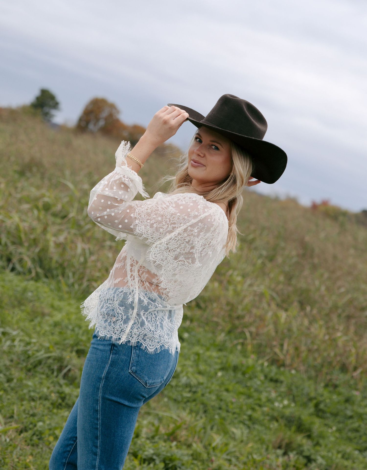 A woman in a white lace top and jeans poses outdoors in a field while wearing a black cowboy hat.