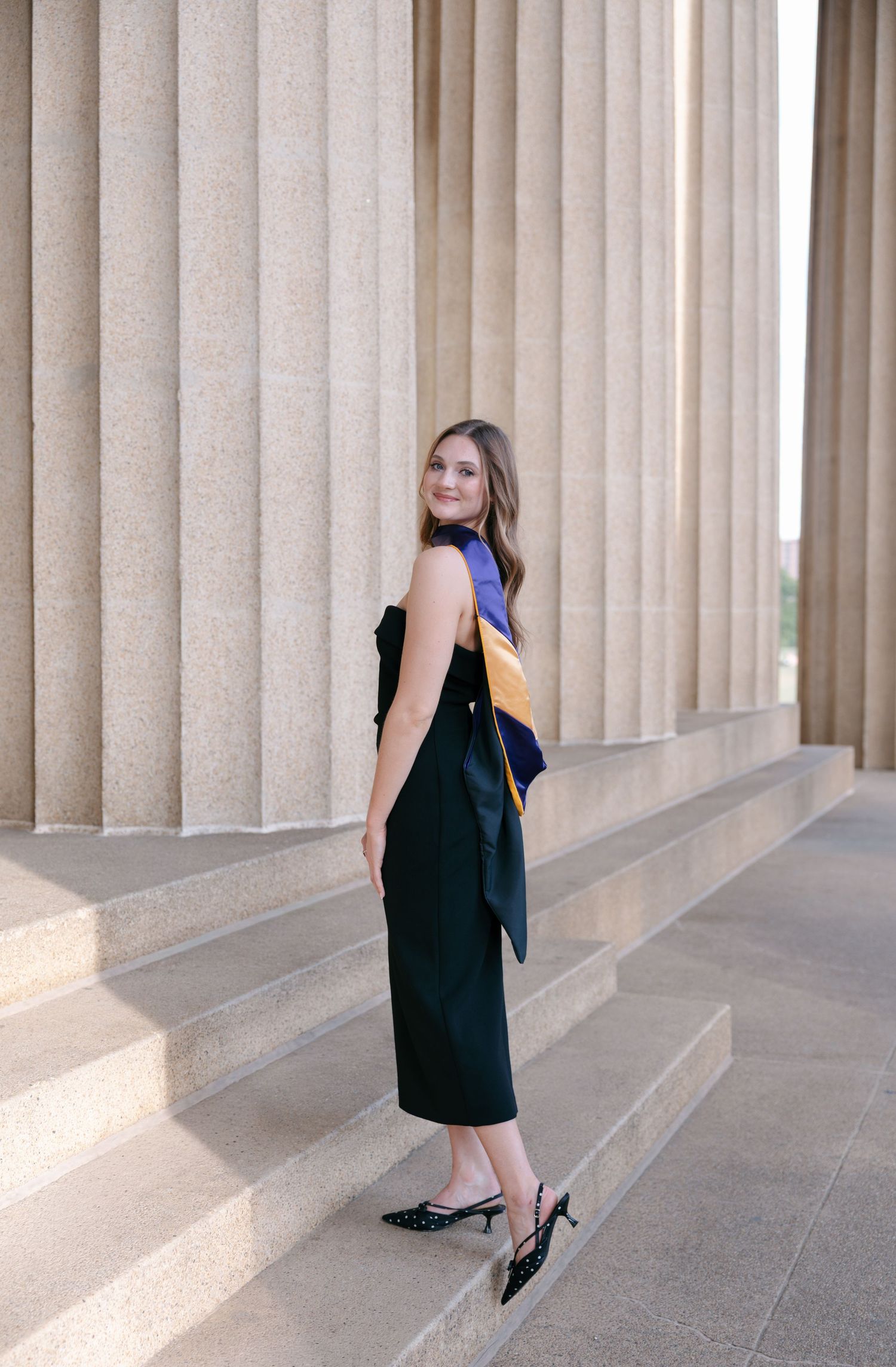 A young woman in a black dress with graduation regalia poses on stone steps beside tall classical columns.