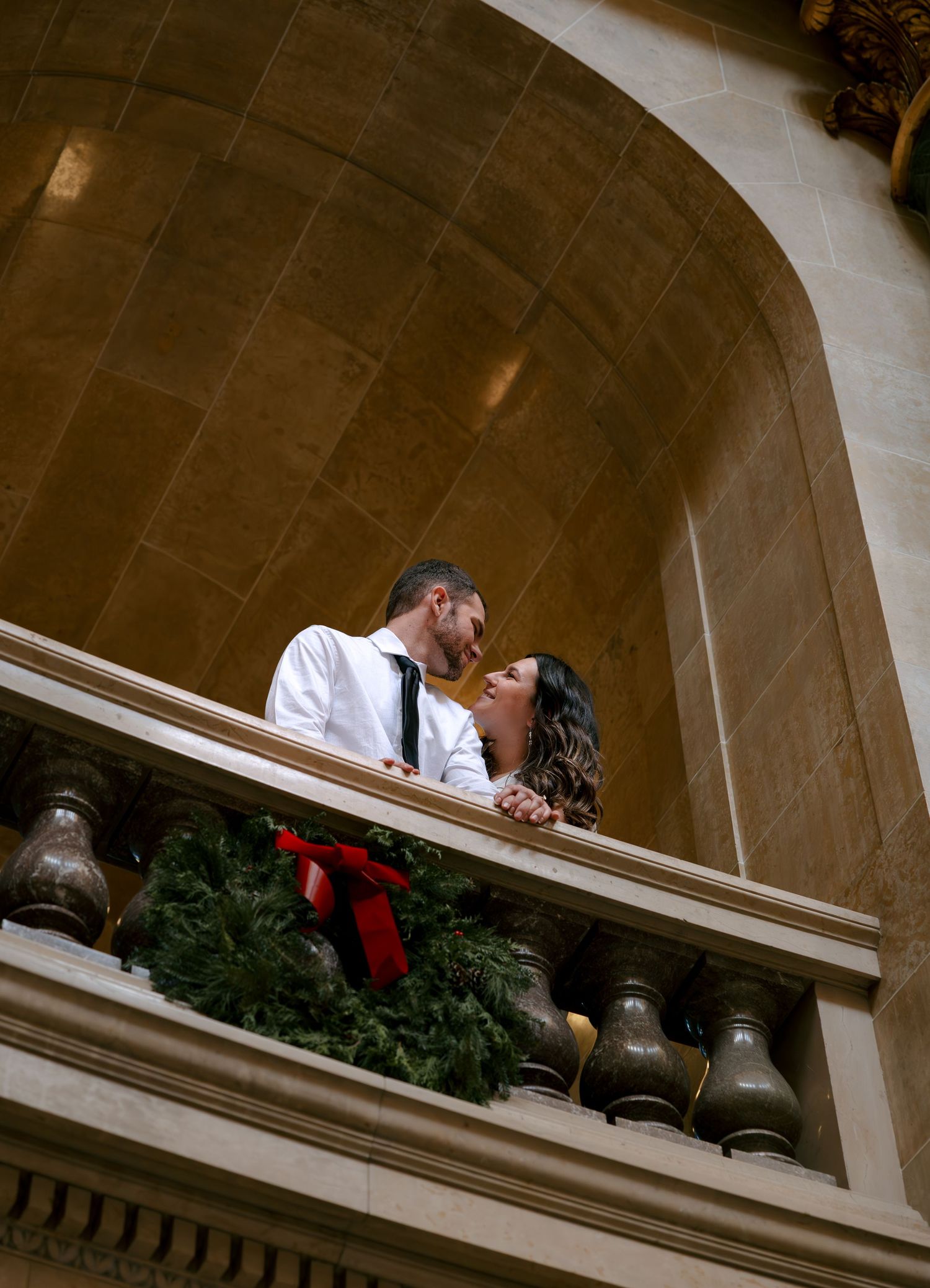 People lean over a stone balcony with Christmas decorations and evergreen garland in an arched architectural setting.