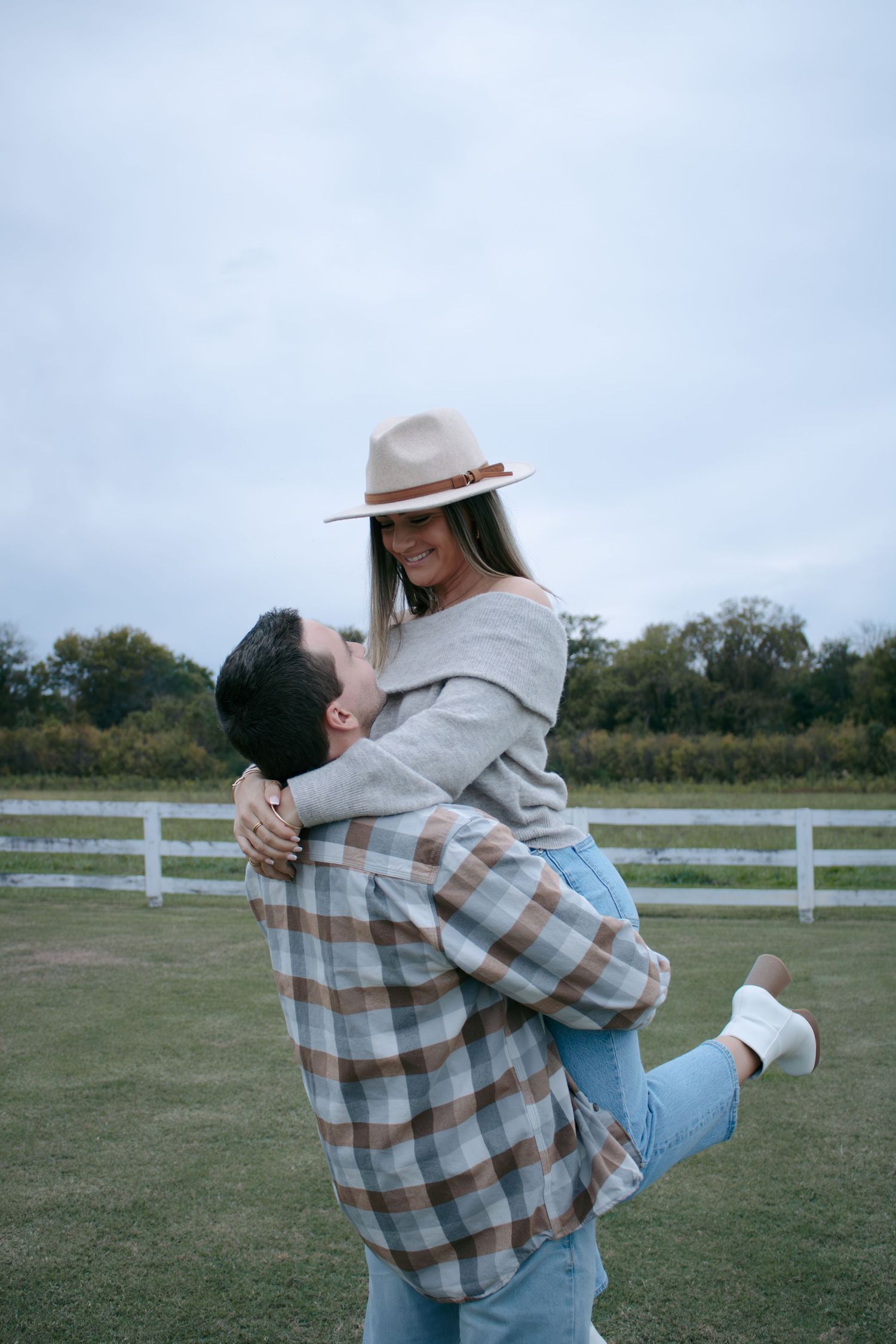 Playful couple embracing and spinning in a field with white fences on a cloudy day.