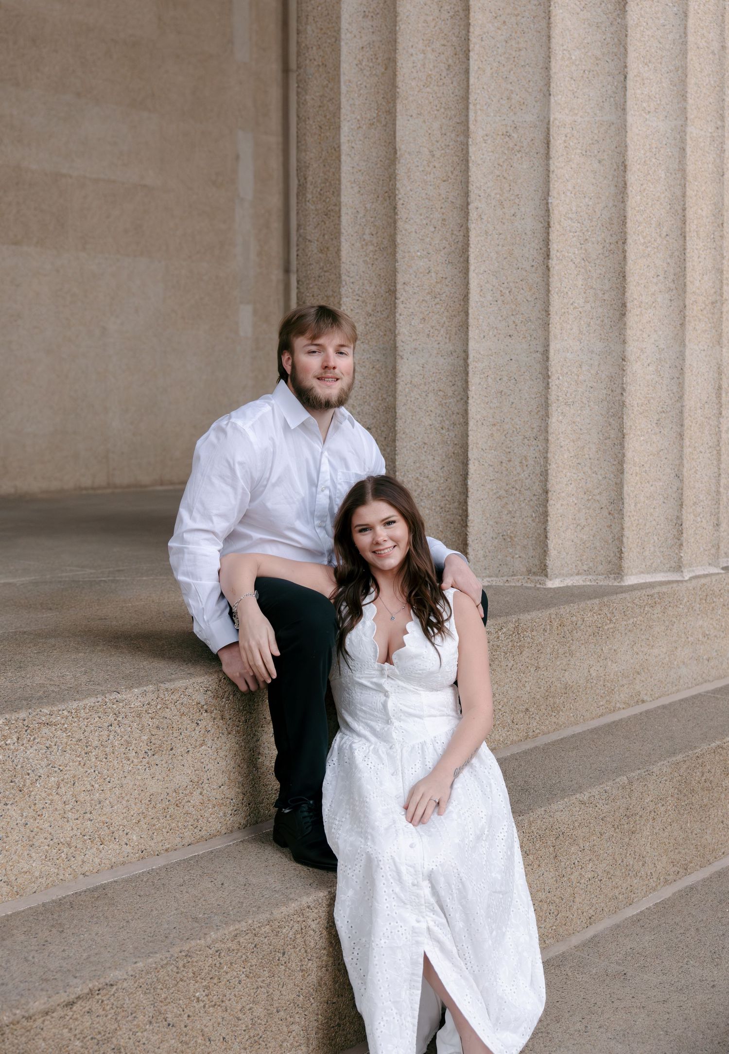 A couple in white outfits poses playfully against stone columns in a series of romantic engagement photos.