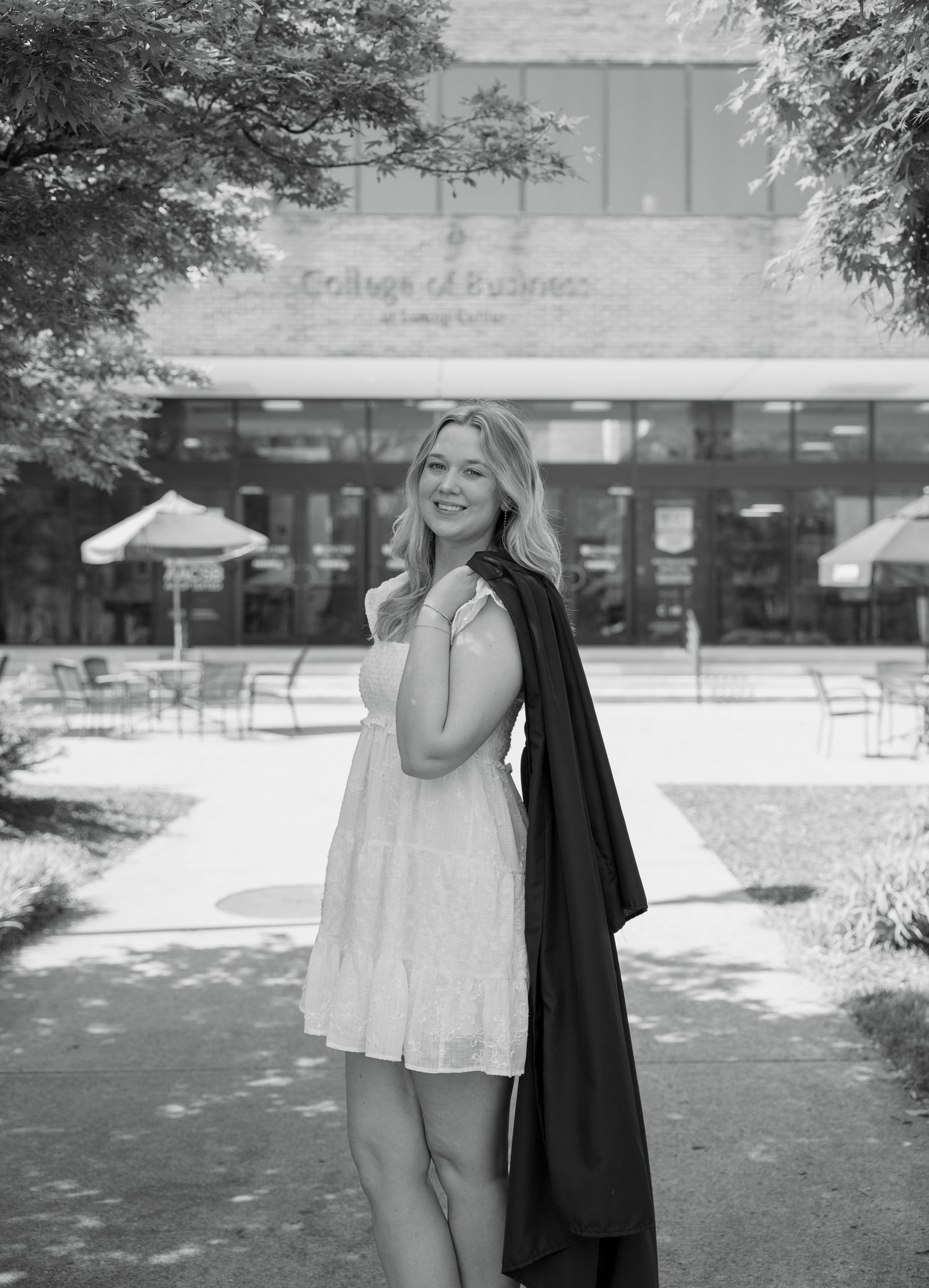 Young woman in white dress holding graduation gown poses outside College of Business building in black and white photo.