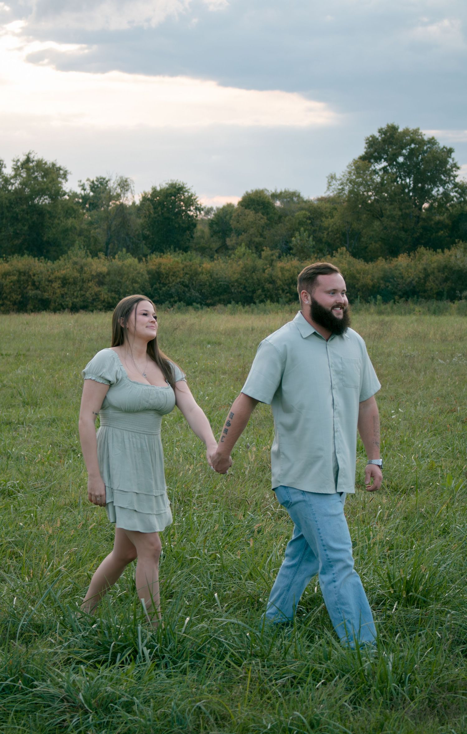 Couple walking hand in hand through tall grass in countryside setting.