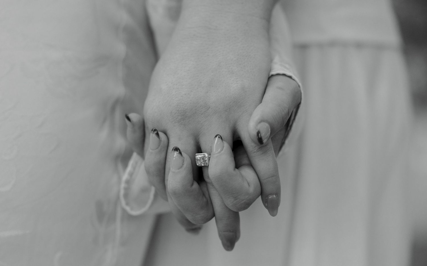 Black and white photo of two people holding hands, one wearing a diamond engagement ring, symbolizing love and commitment.
