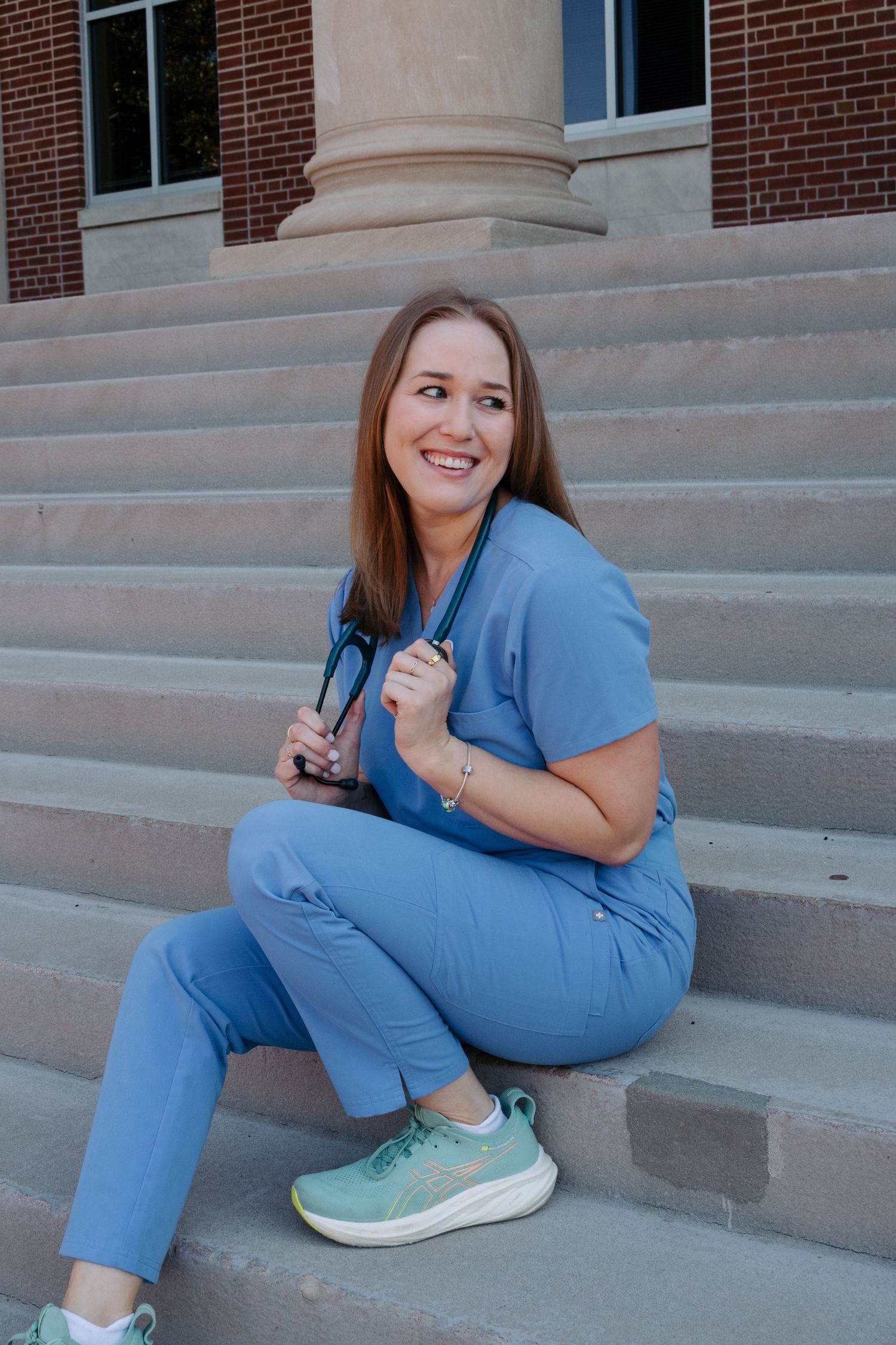 A person in blue scrubs and mint green sneakers sits and poses on concrete steps outside a brick building.