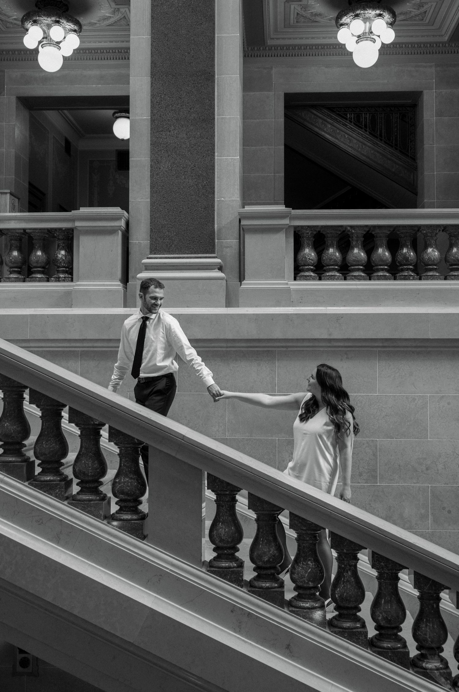 A person in white dress descends an elegant marble staircase with ornate black railings in a classic building interior.