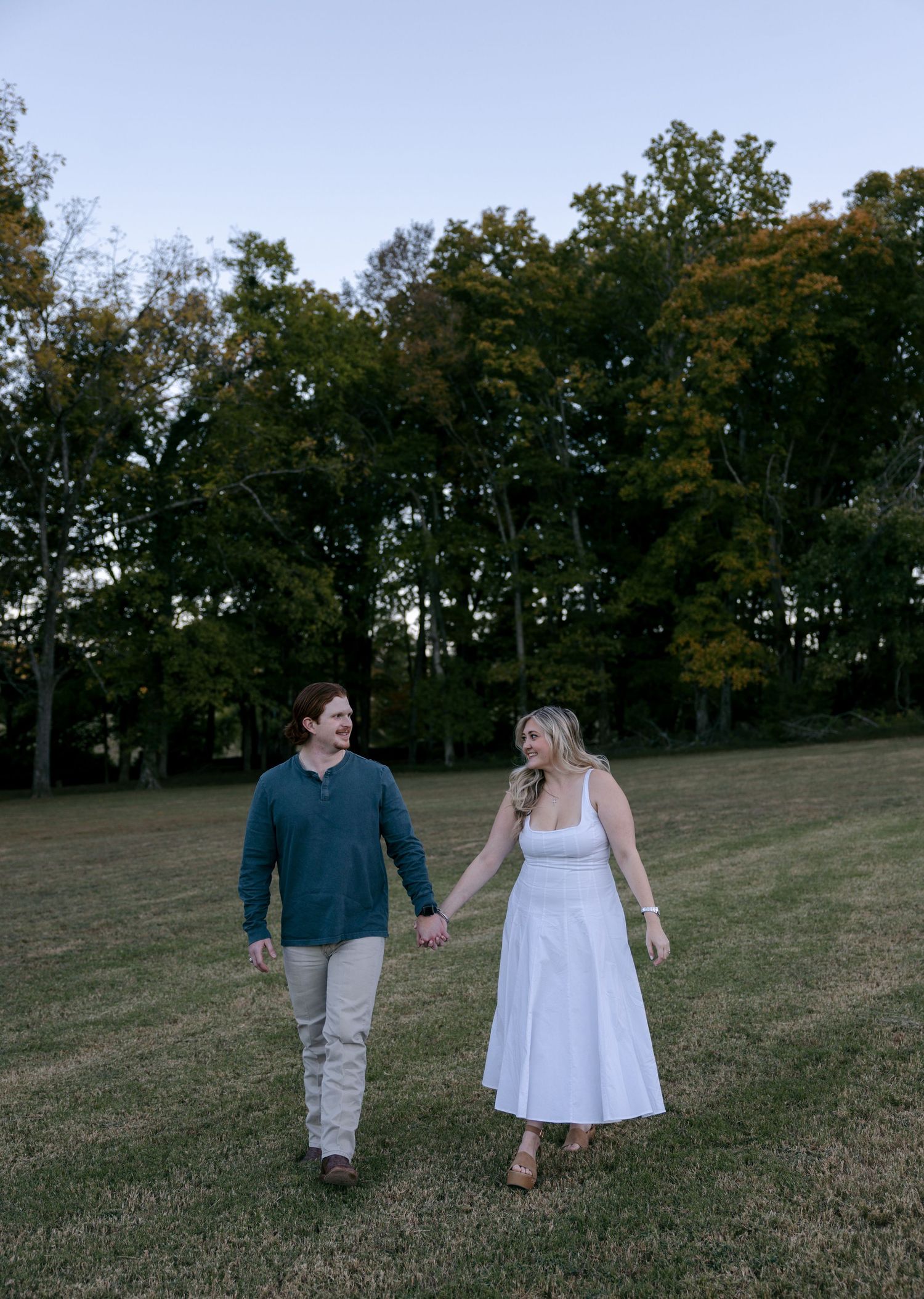 Smiling couple holding hands and walking across a grassy field with autumn trees behind them, he in a teal sweater and she in a white dress.