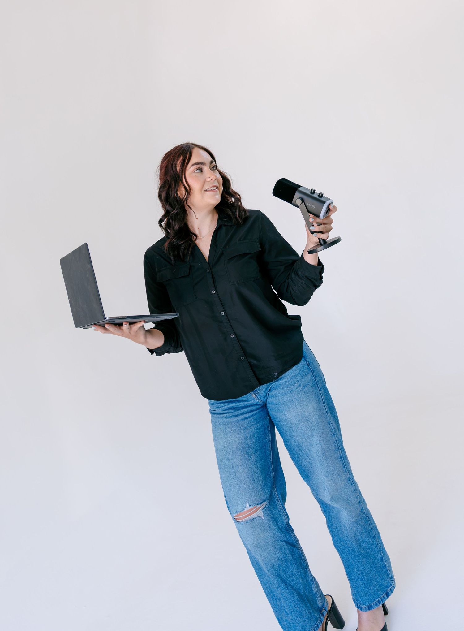 Woman in black shirt and jeans holds laptop and microphone, smiling confidently against white background.