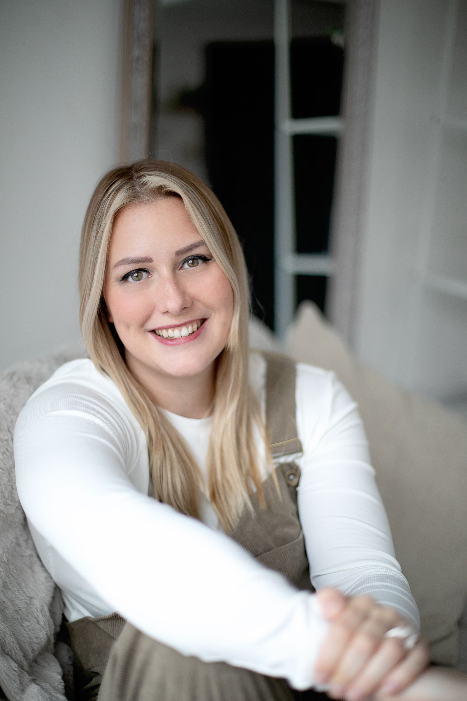 Portrait of woman in white sweater and beige overalls sitting on grey couch. Ephemera.