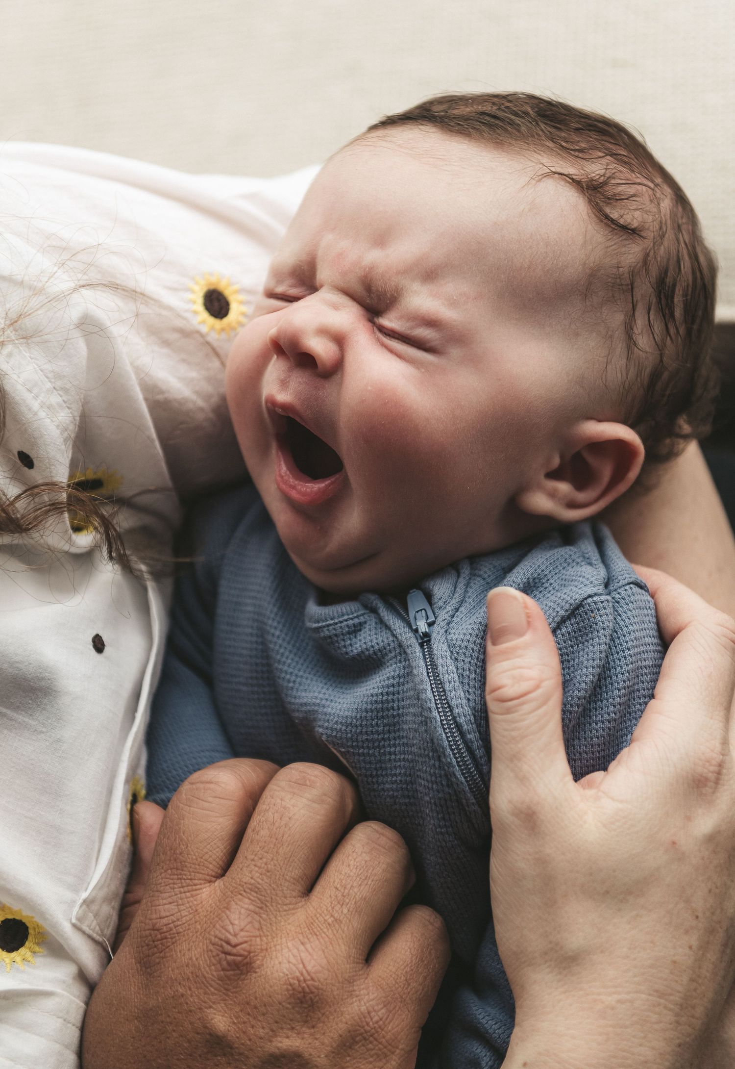 A yawning baby wearing a blue outfit lies on a white floral patterned blanket. Ephemera.