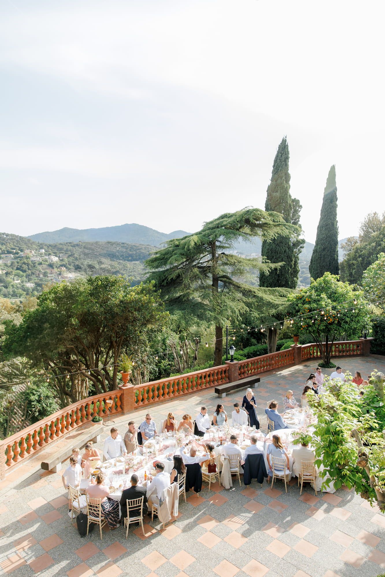 Elegant outdoor wedding reception on a terraced villa patio with mountain views, lush cypress trees, and guests dining al fresco.