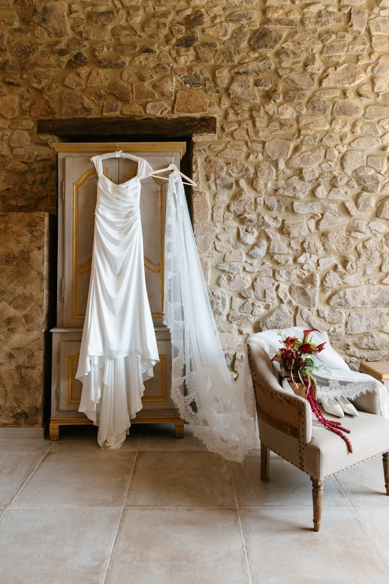 White wedding dress hanging on rustic wooden armoire with lace veil beside bridal bouquet on vintage chair against stone wall.