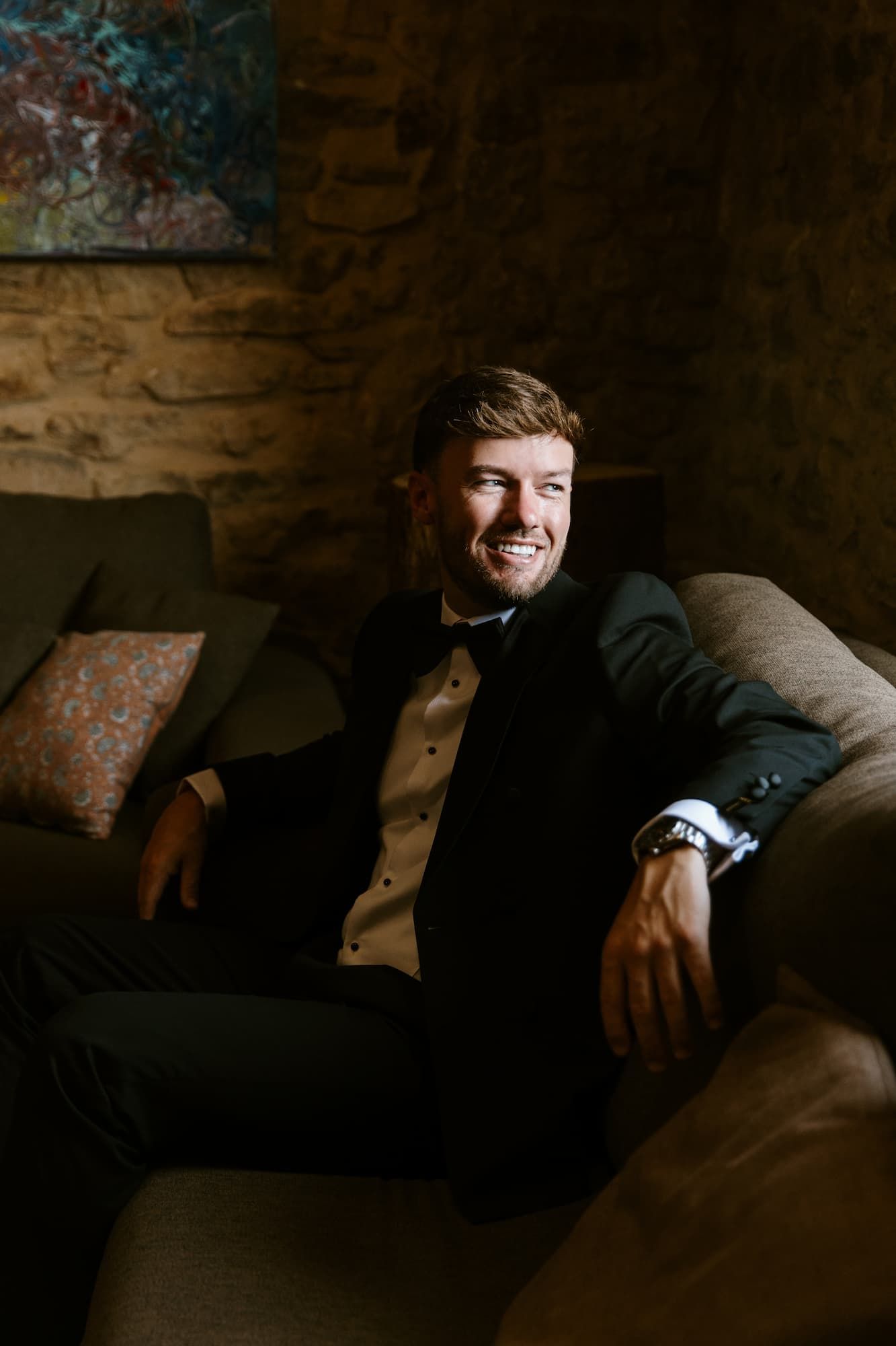 Smiling groom in black tuxedo and bow tie relaxing on a sofa in a rustic stone-walled venue.