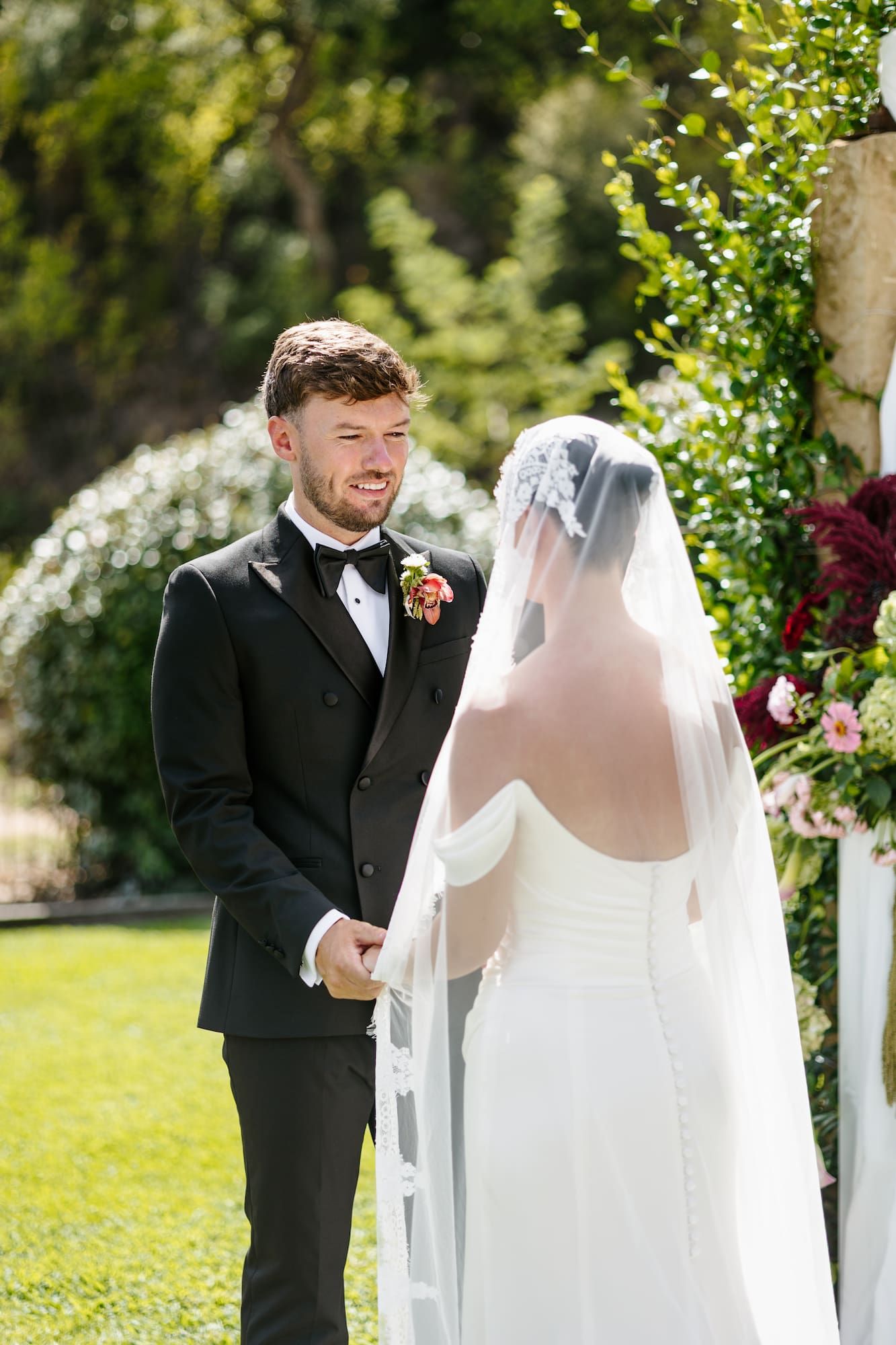 Groom in black tuxedo smiling at bride in white strapless gown and veil during outdoor wedding ceremony.