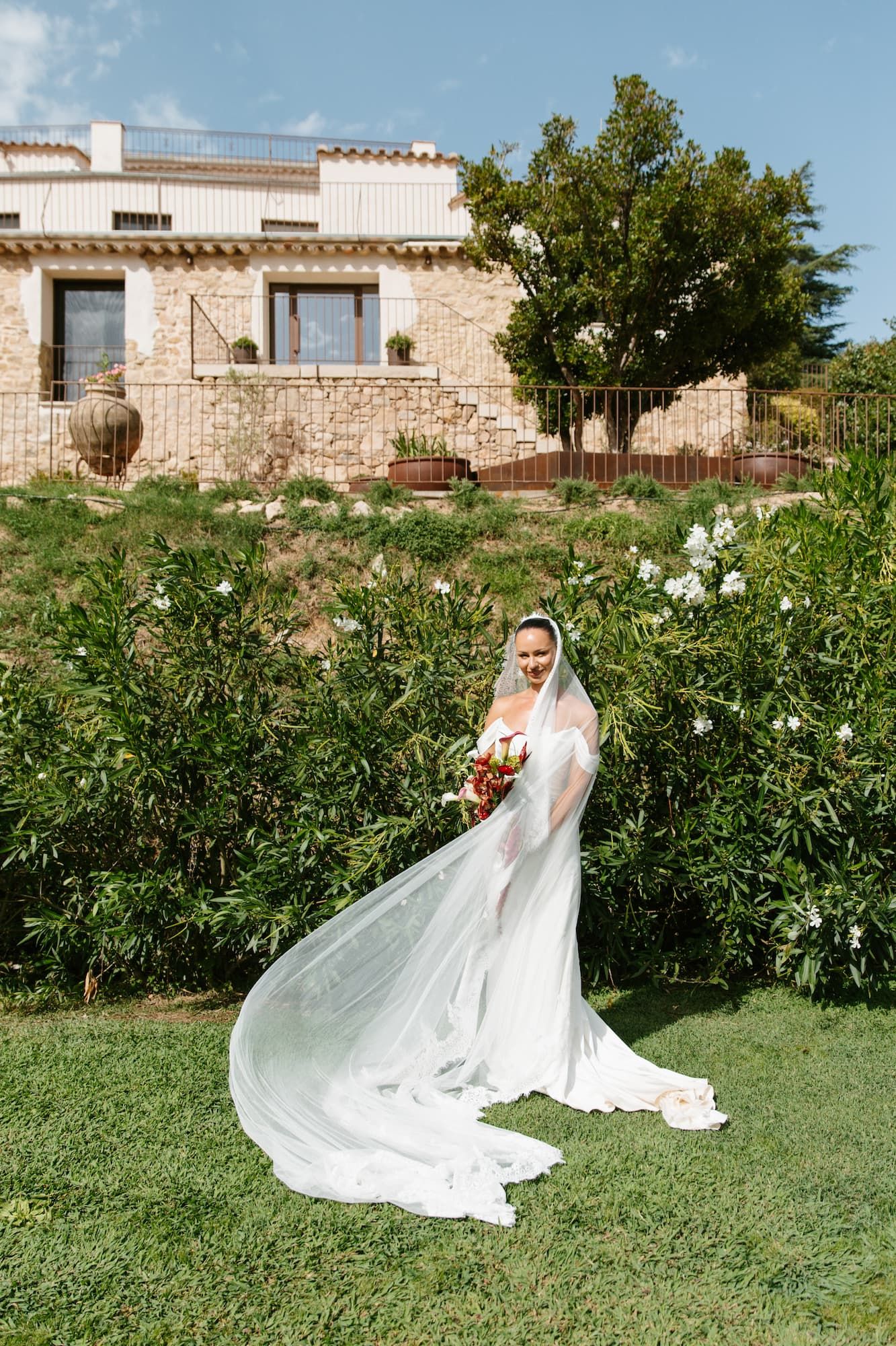 Bride in white gown and veil holding bouquet stands on green lawn before a stone villa on a sunny day.