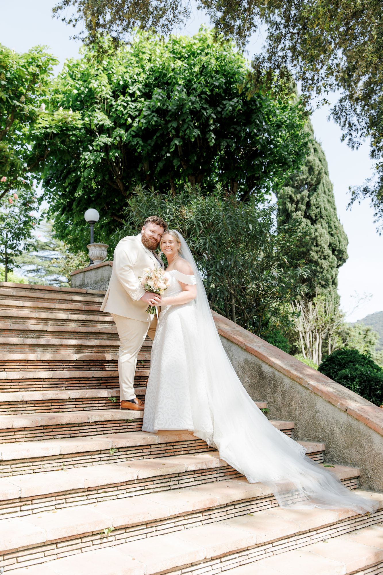 Bride and groom pose on stone steps surrounded by lush greenery on their wedding day.