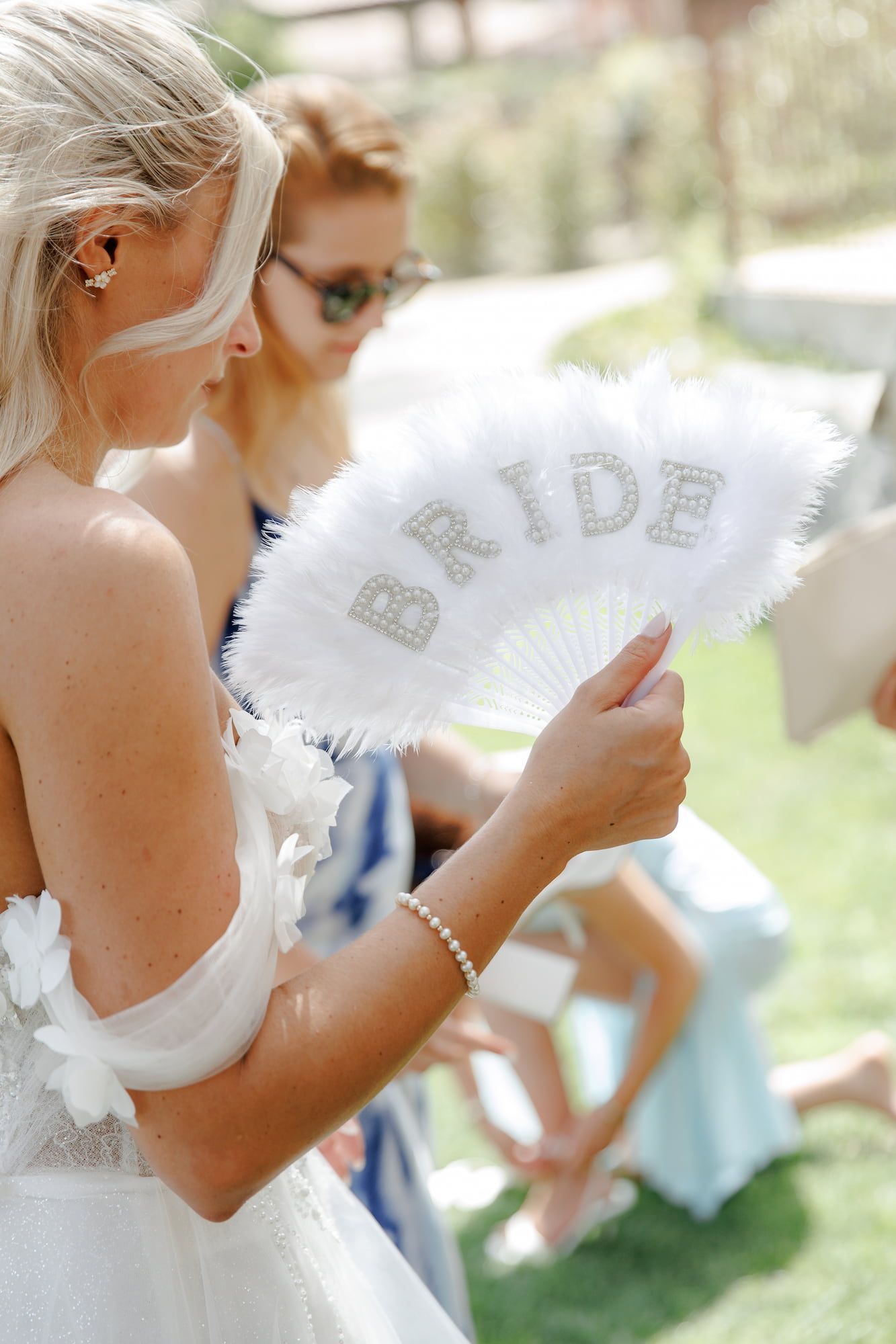 Bride holding white feather fan with rhinestone BRIDE text at outdoor wedding ceremony near Barcelona