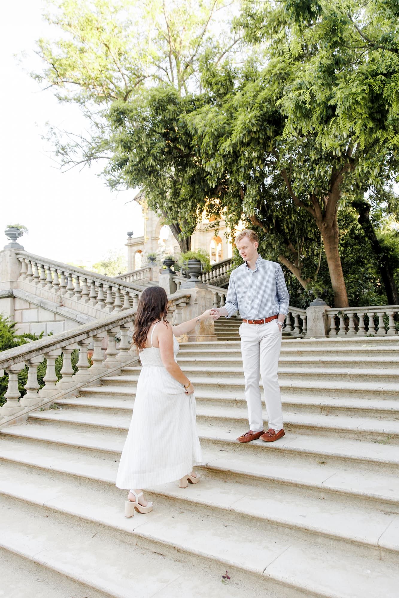 Couple holding hands on elegant stone staircase surrounded by lush green trees, woman in white dress, man in light blue shirt.