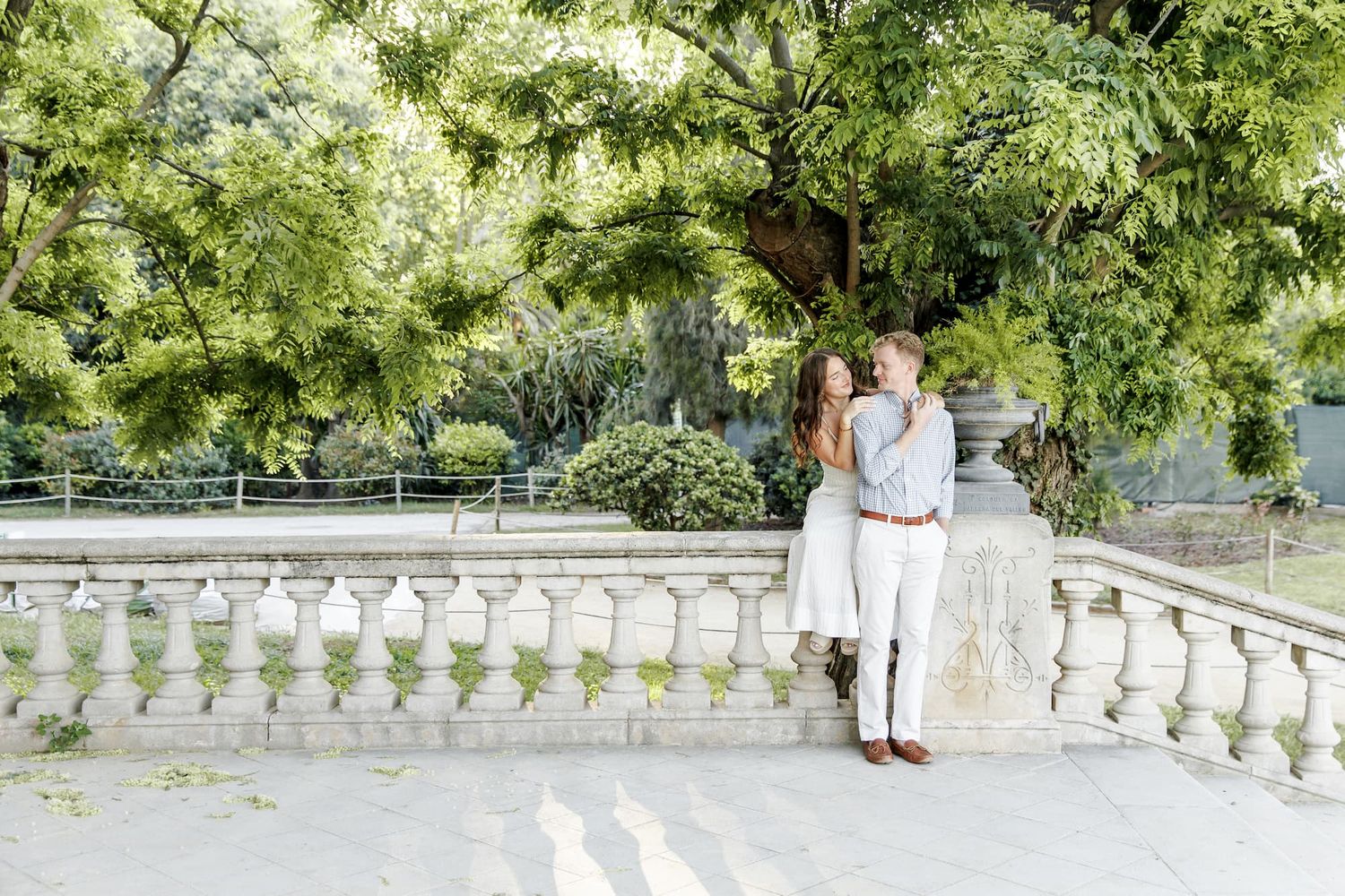 Couple embracing on a white stone balustrade terrace surrounded by lush green trees in an elegant outdoor setting.