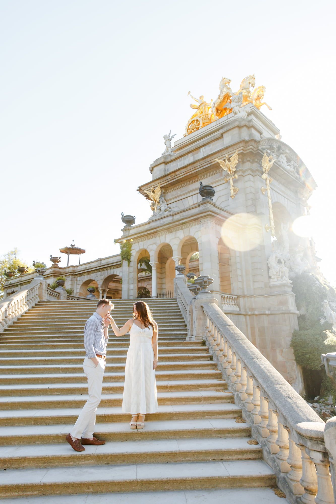 Couple holding hands on grand baroque staircase with golden statues and sunlight flare in background.