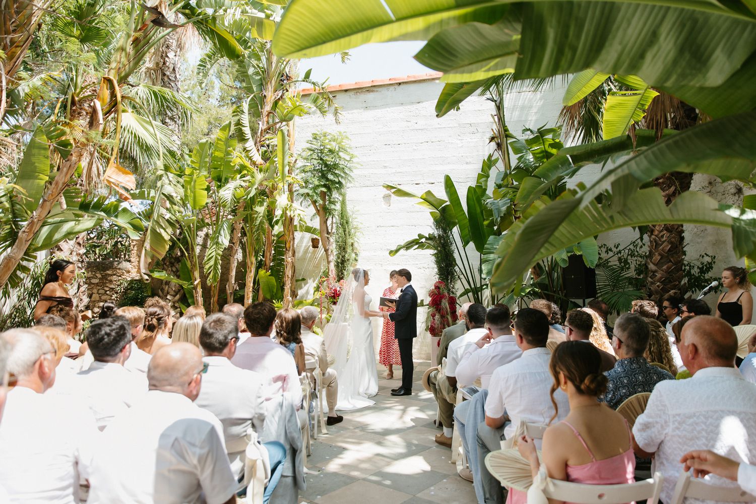 Outdoor wedding ceremony surrounded by lush tropical plants with guests seated in white chairs.