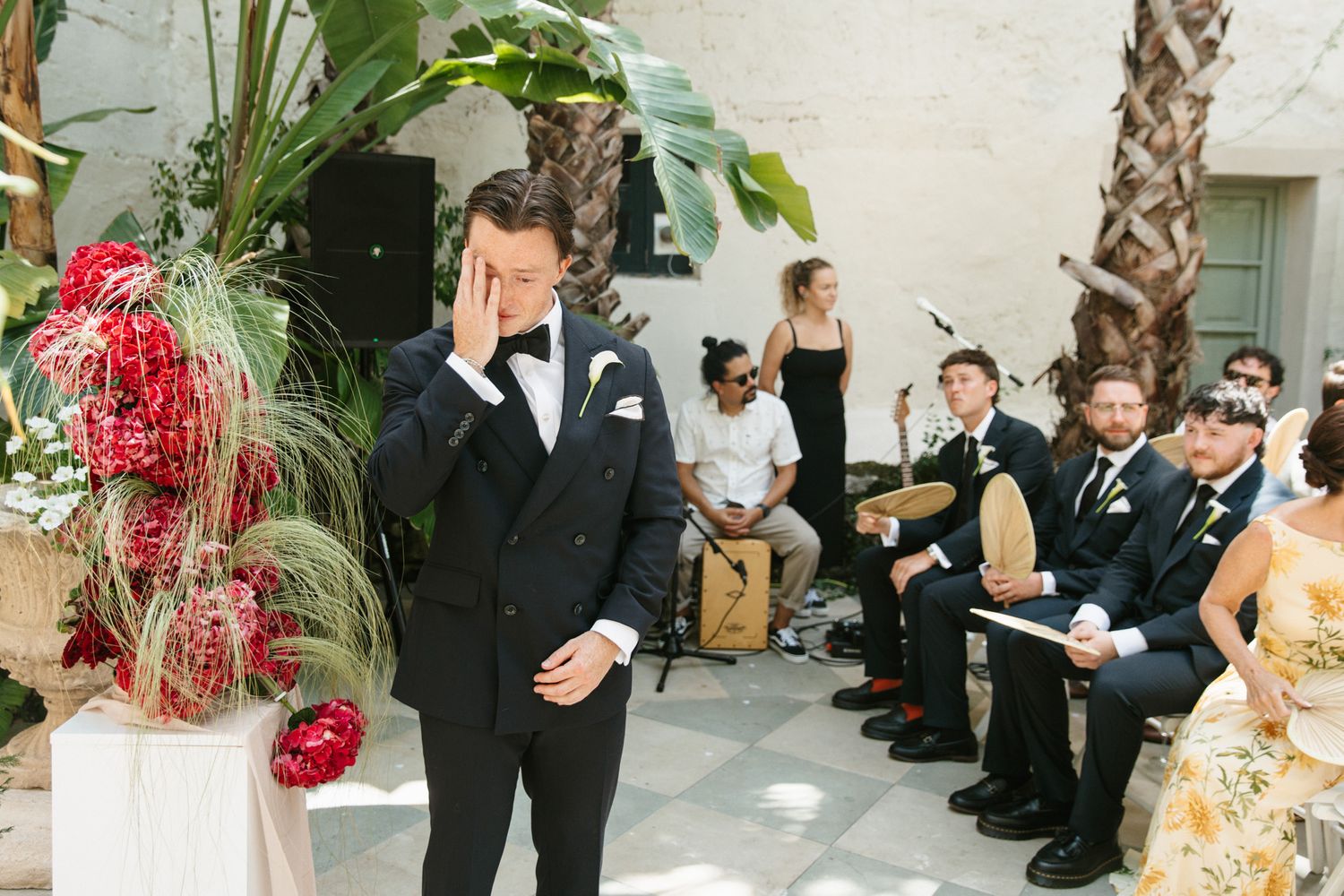 Emotional groom in black tuxedo wiping tears at outdoor wedding ceremony with live band and tropical floral arrangements.