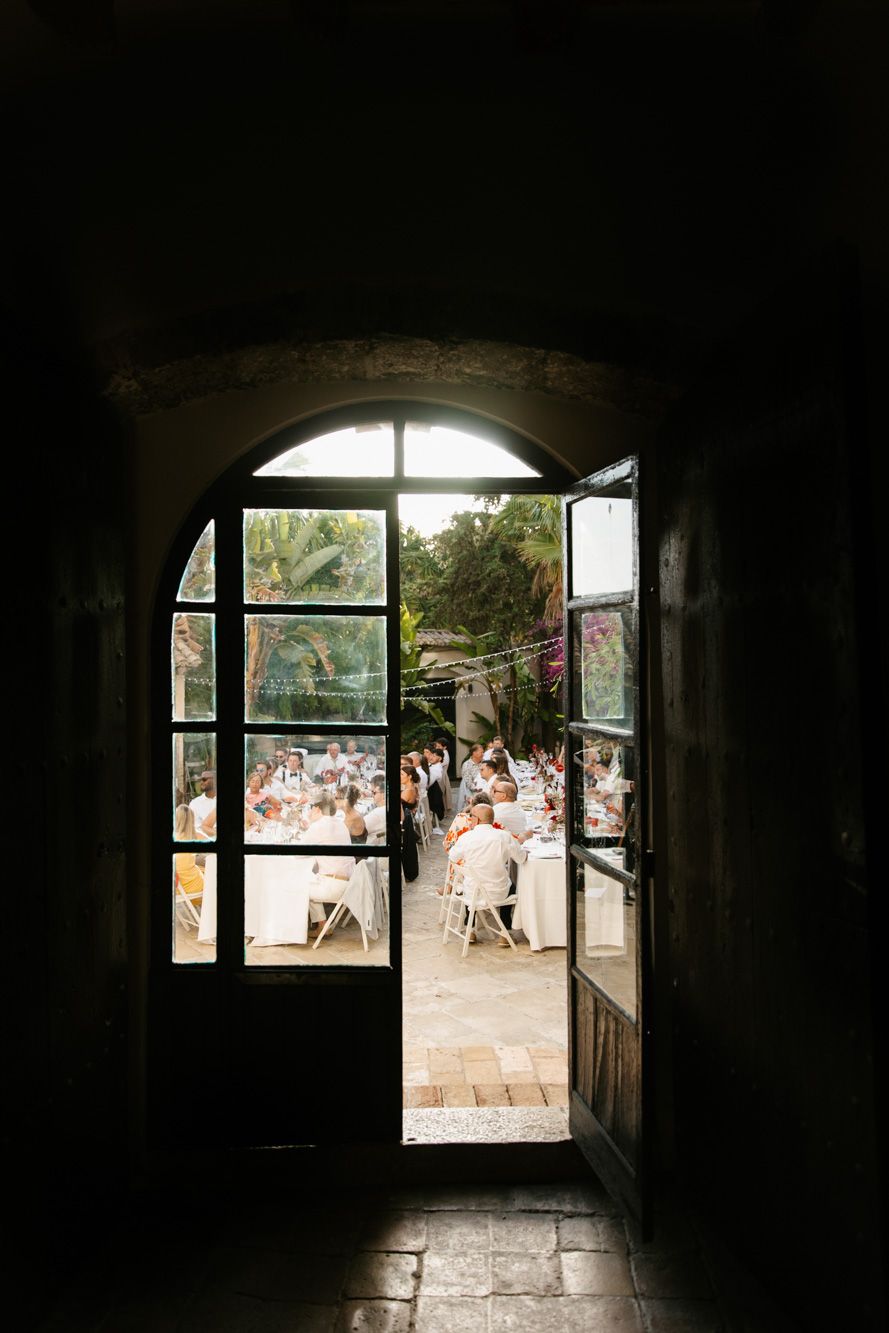 Dark arched doorway opening to a sunlit outdoor wedding reception with guests seated at white tables amid lush greenery.