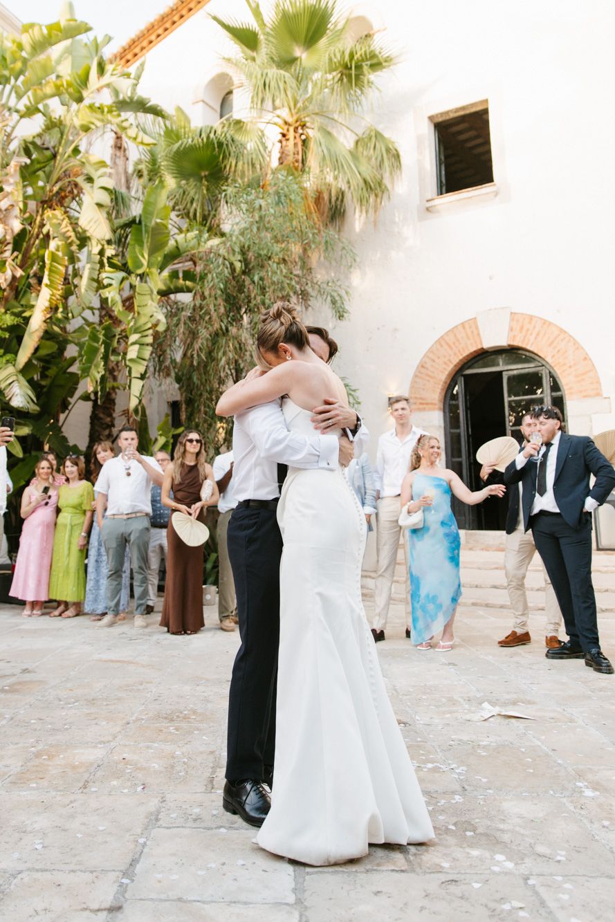 Bride and groom share first dance outdoors at Mediterranean villa wedding, surrounded by guests and palm trees.