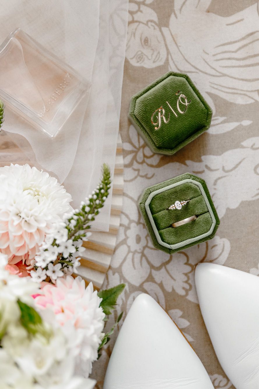 Green velvet monogrammed ring box with diamond engagement ring beside bridal bouquet and white heels on floral fabric.