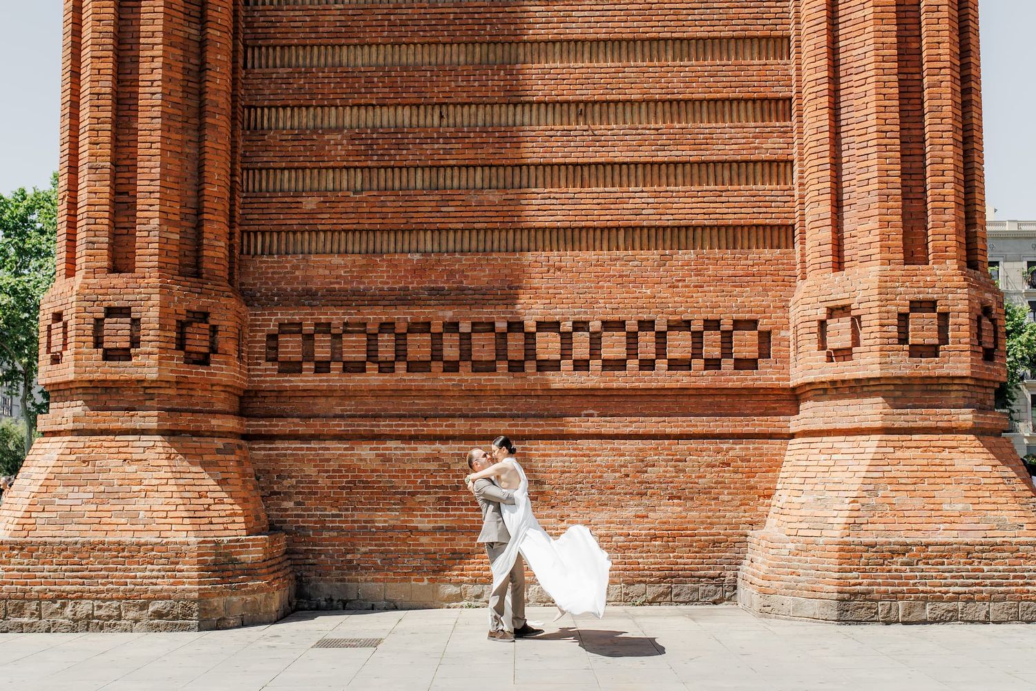 A white wedding dress flows elegantly against the red brick of the Arc de Triomphe in Barcelona