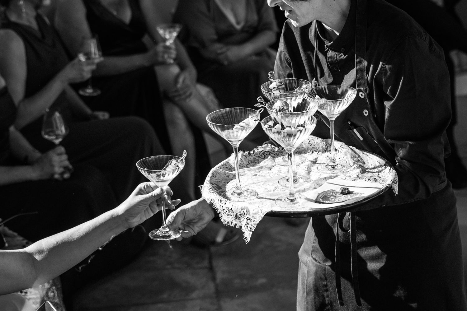 Black and white photo of a waiter holding a tray of stacked champagne coupe glasses while a guest reaches for a drink at an elegant event.