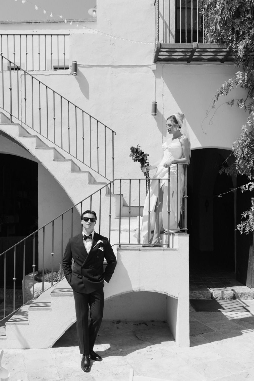 Black and white wedding photo of groom in tuxedo standing below bride in white dress on Mediterranean staircase.