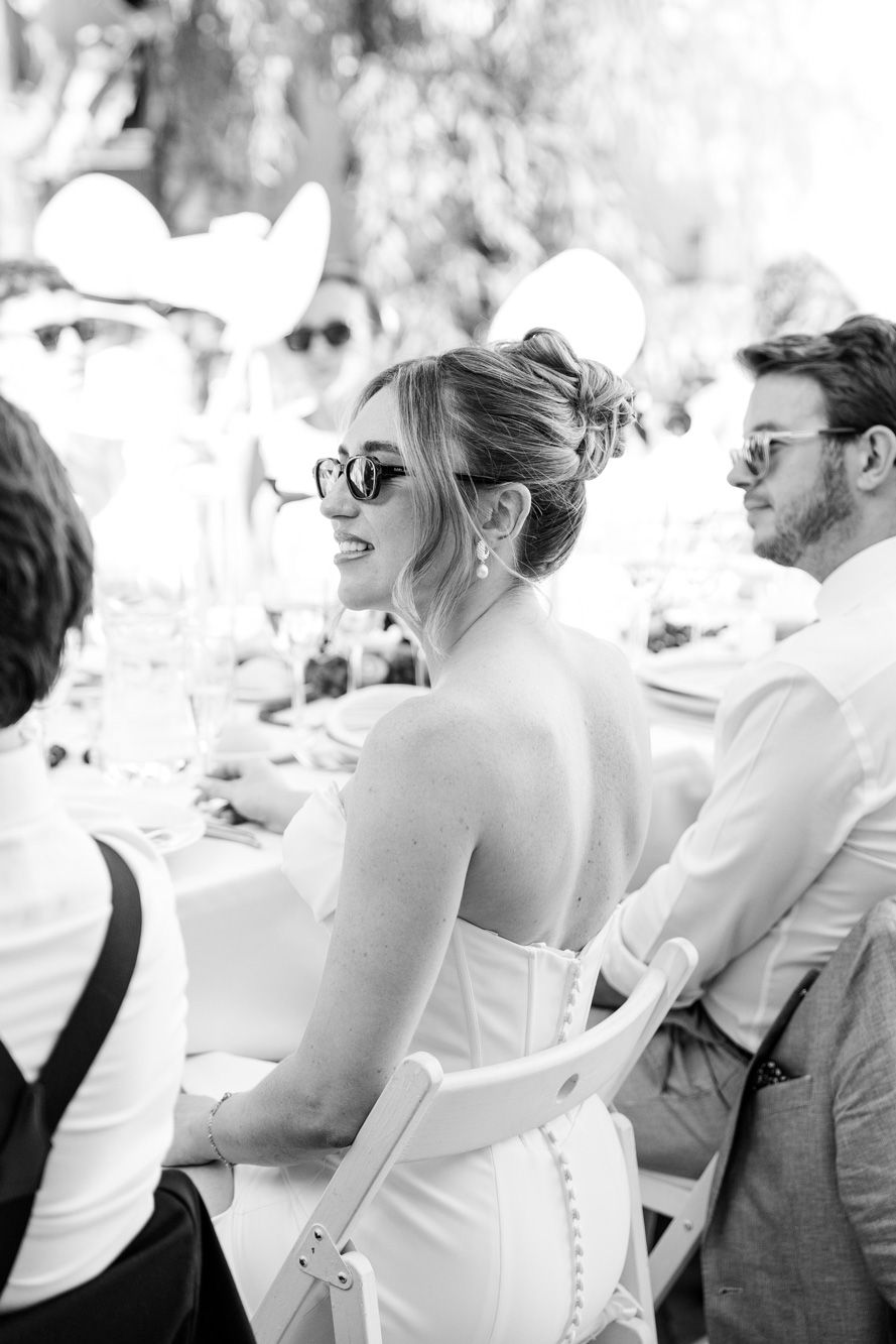 Bride in white strapless gown wearing sunglasses smiles at outdoor wedding reception table.