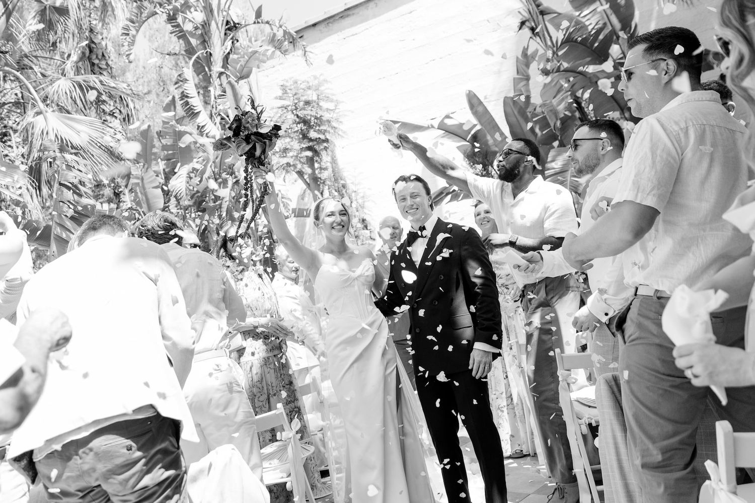 Black and white photo of a joyful bride and groom walking through confetti after their wedding ceremony.