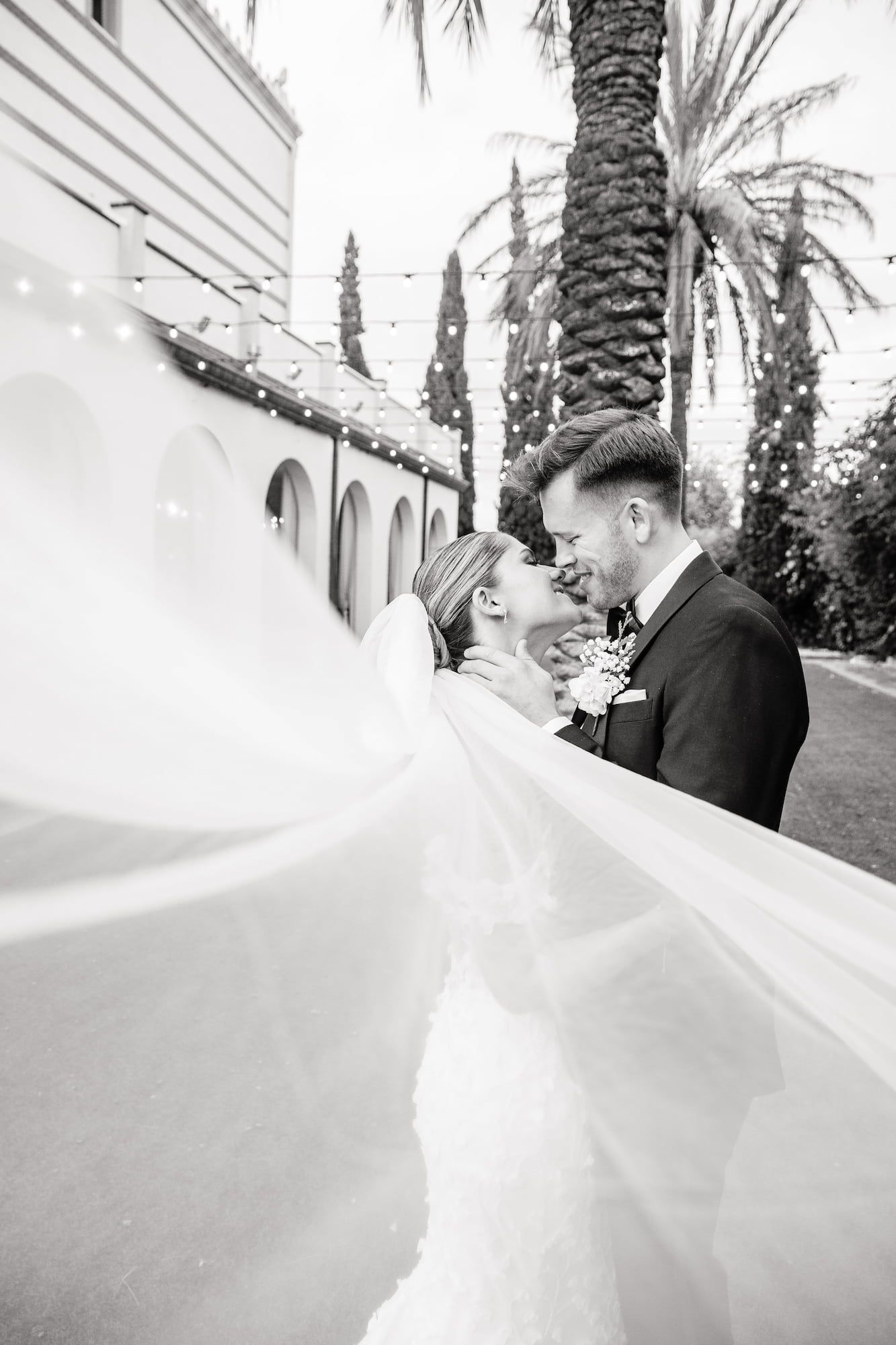 Black and white wedding photo of bride and groom embracing, veil flowing, beside a white building with palm tree backdrop.