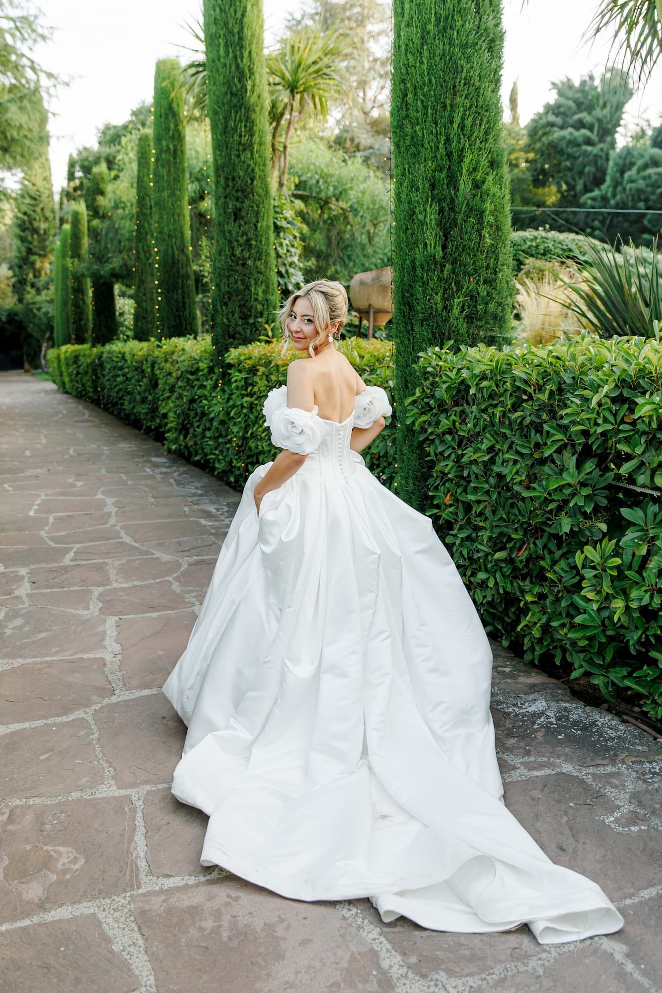 Bride in elegant off-shoulder white gown with long train walks through garden at the wedding venue Gran Villa Rosa in Barcelona