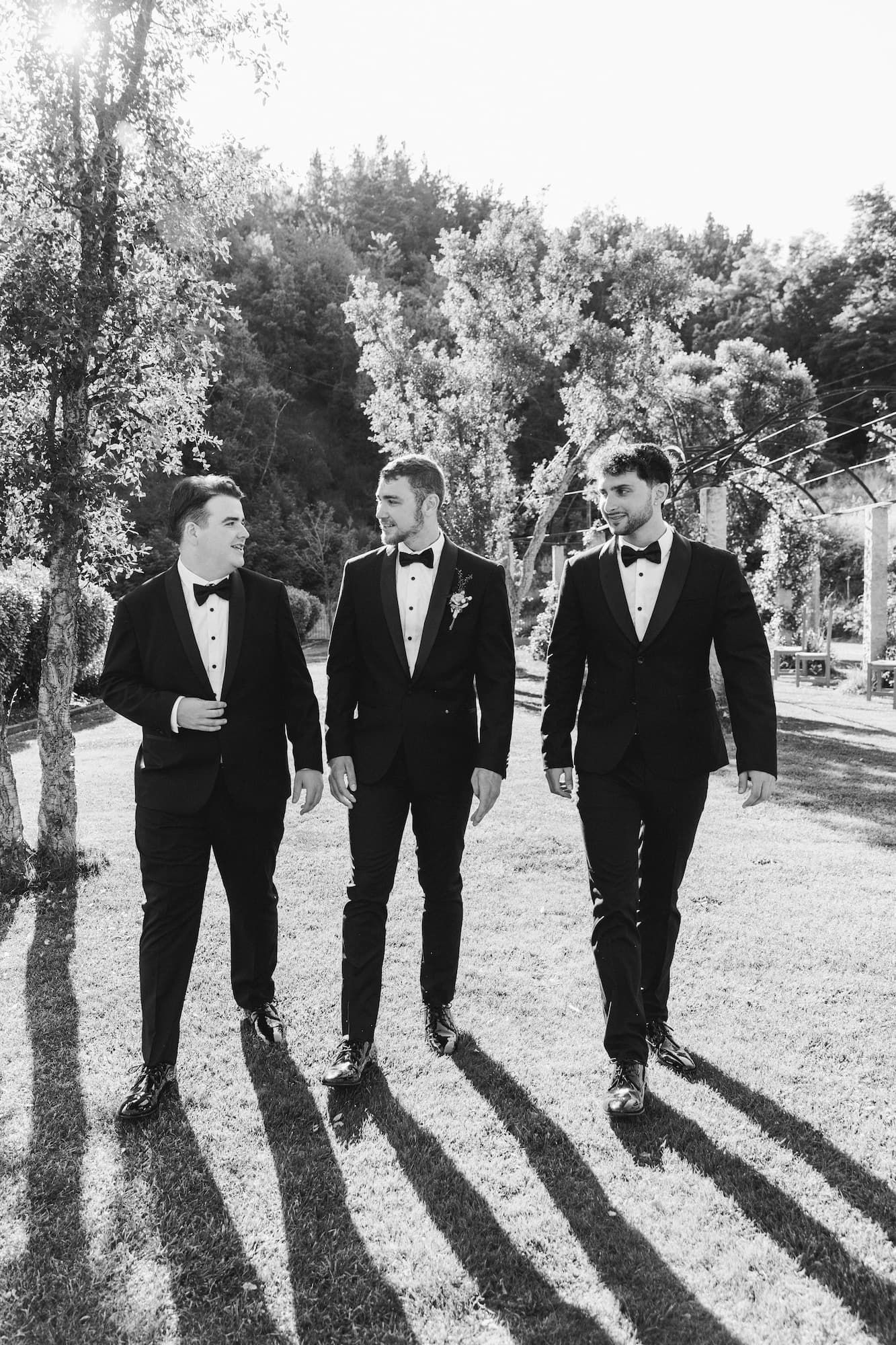 A wedding party in black suits walks across a grassy lawn, casting long shadows in dramatic black-and-white photography.
