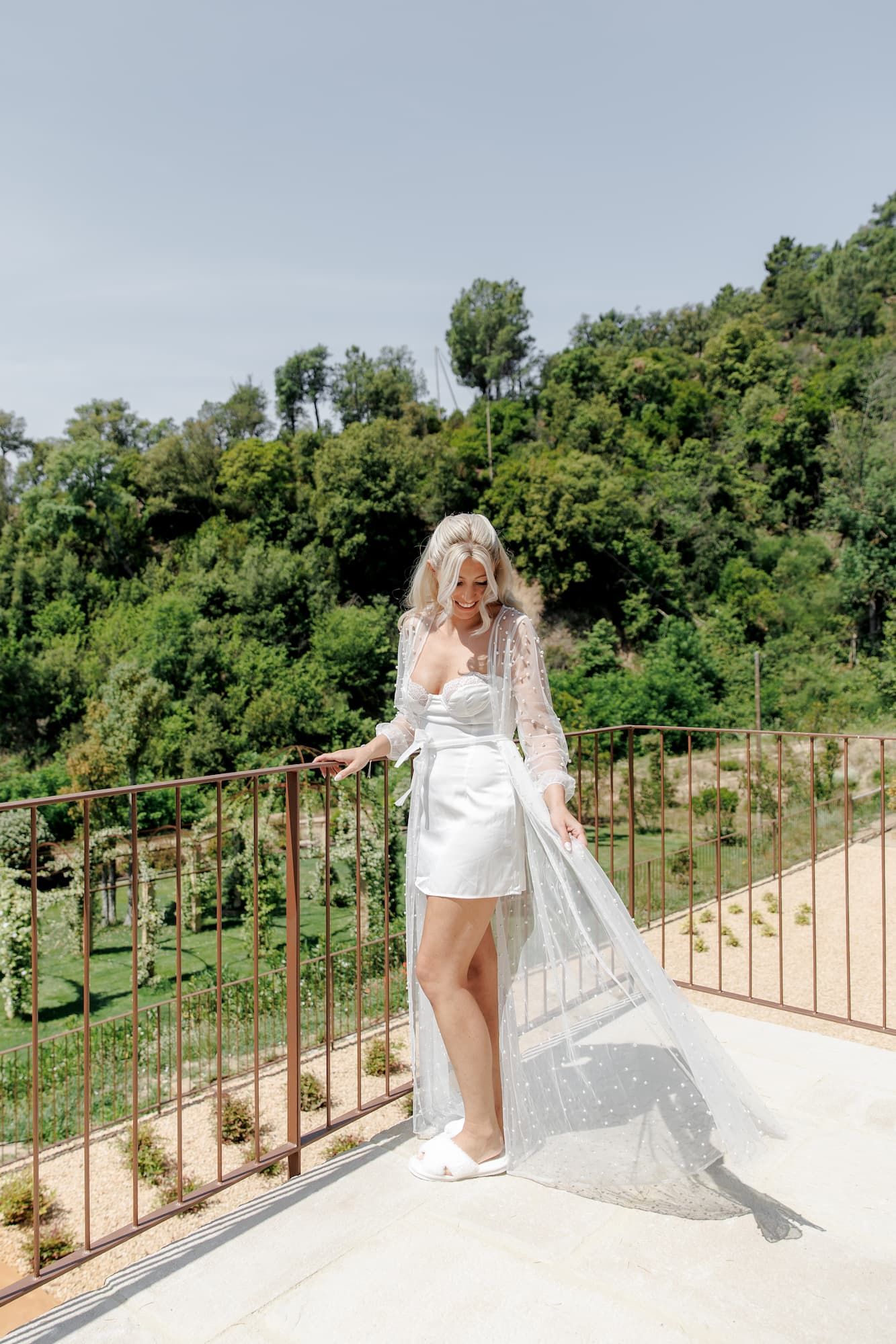 Bride in short white dress and veil stands on balcony surrounded by lush green trees on sunny day.